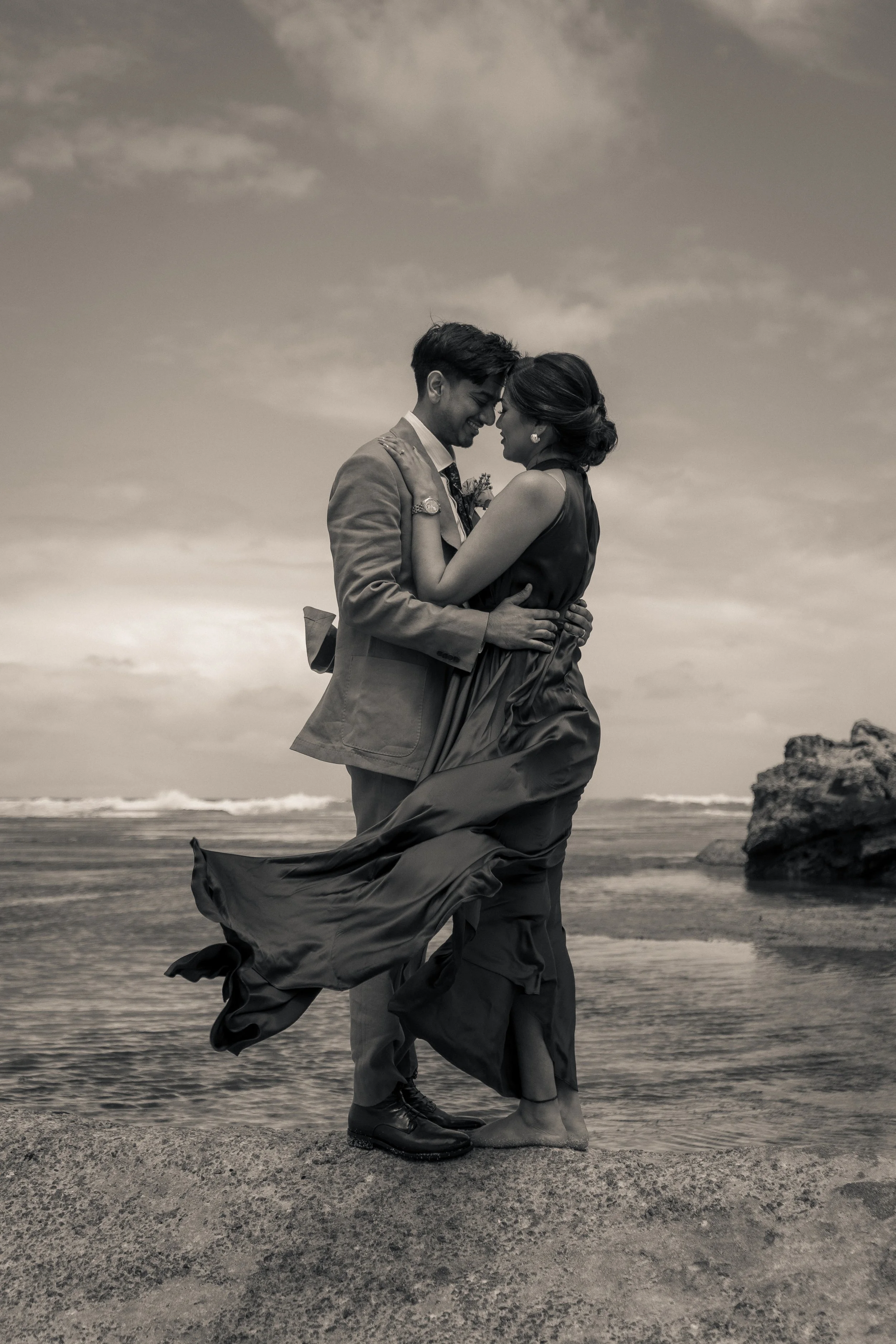 A black and white wedding photo of a couple embracing on a beach, with the man in a suit and the woman in a flowing dress, their foreheads touching, ocean waves in the background. Beach wedding/elopement