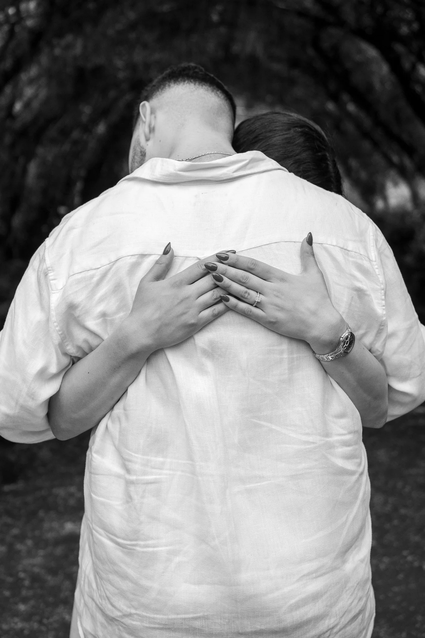A person wearing a white shirt and a woman with a watch and wedding band hugging from behind, with their faces hidden.