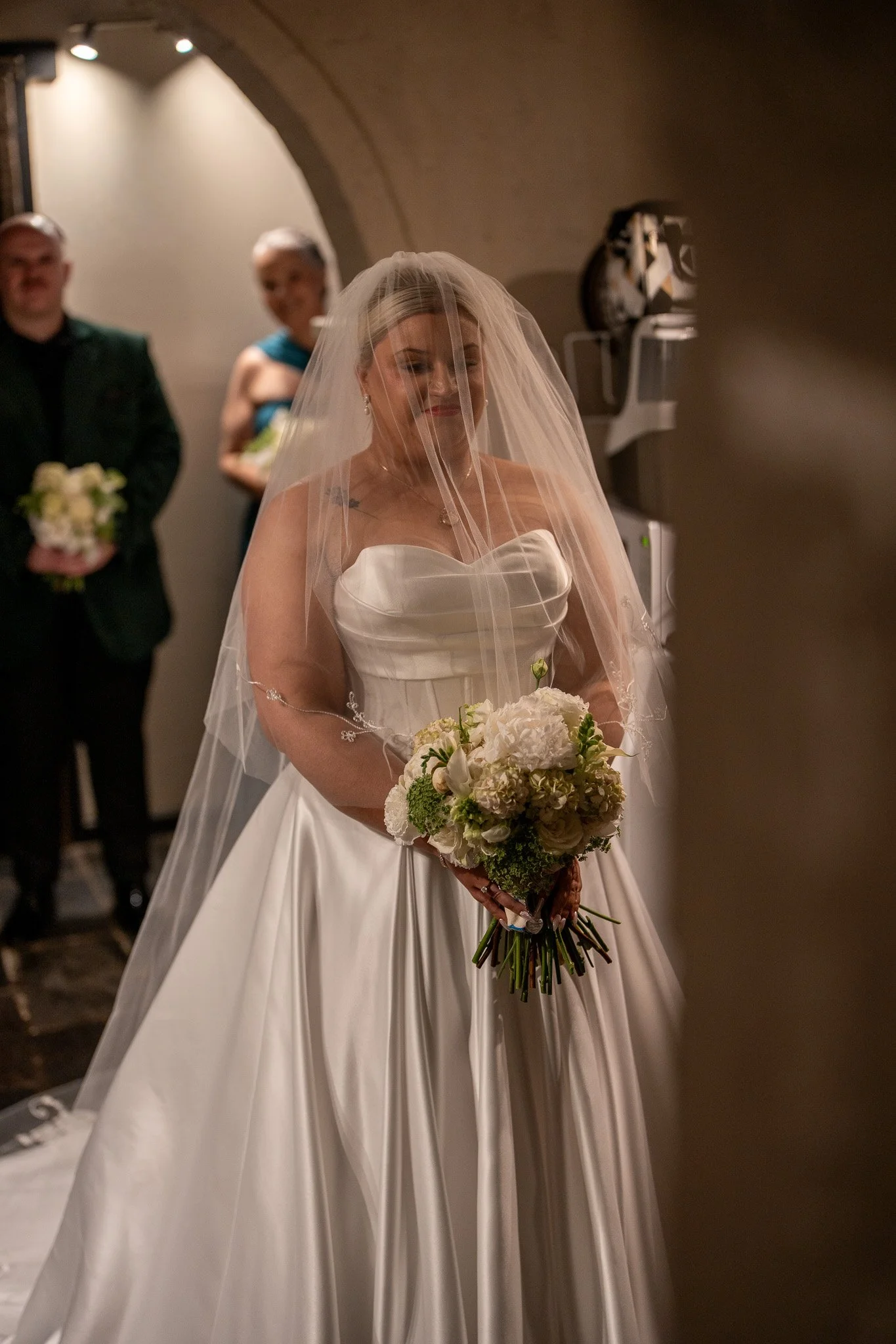 A bride in a white wedding dress with a veil, holding a bouquet of white and green flowers, smiling with eyes closed during a wedding ceremony.