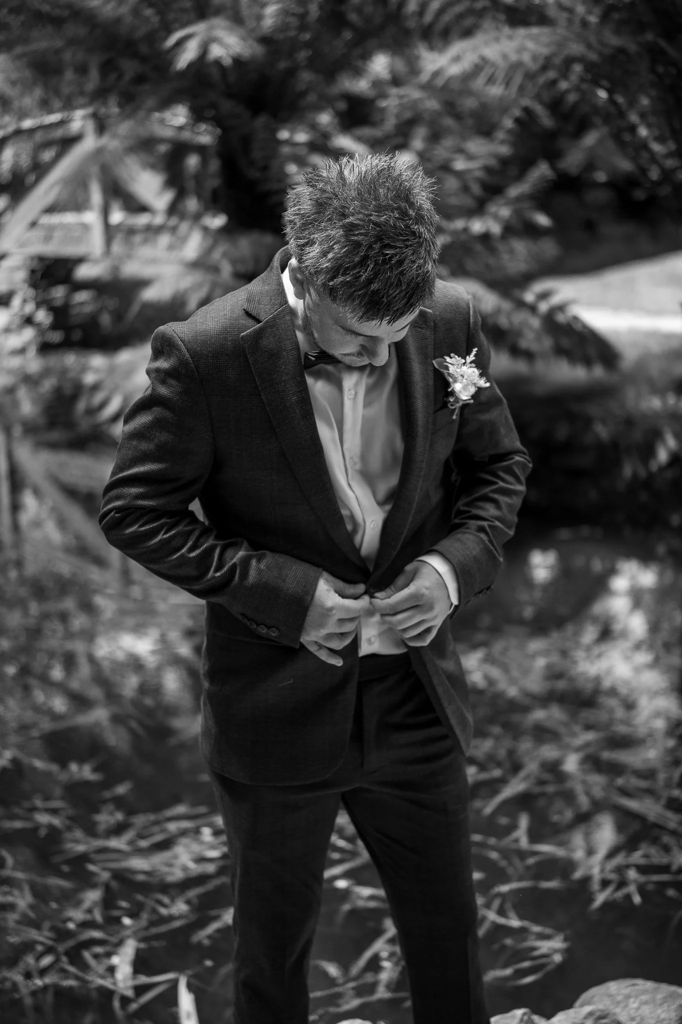 A man dressed in a suit and bowtie, standing outdoors, buttoning his jacket, with a boutonniere on his lapel, near a body of water and trees.