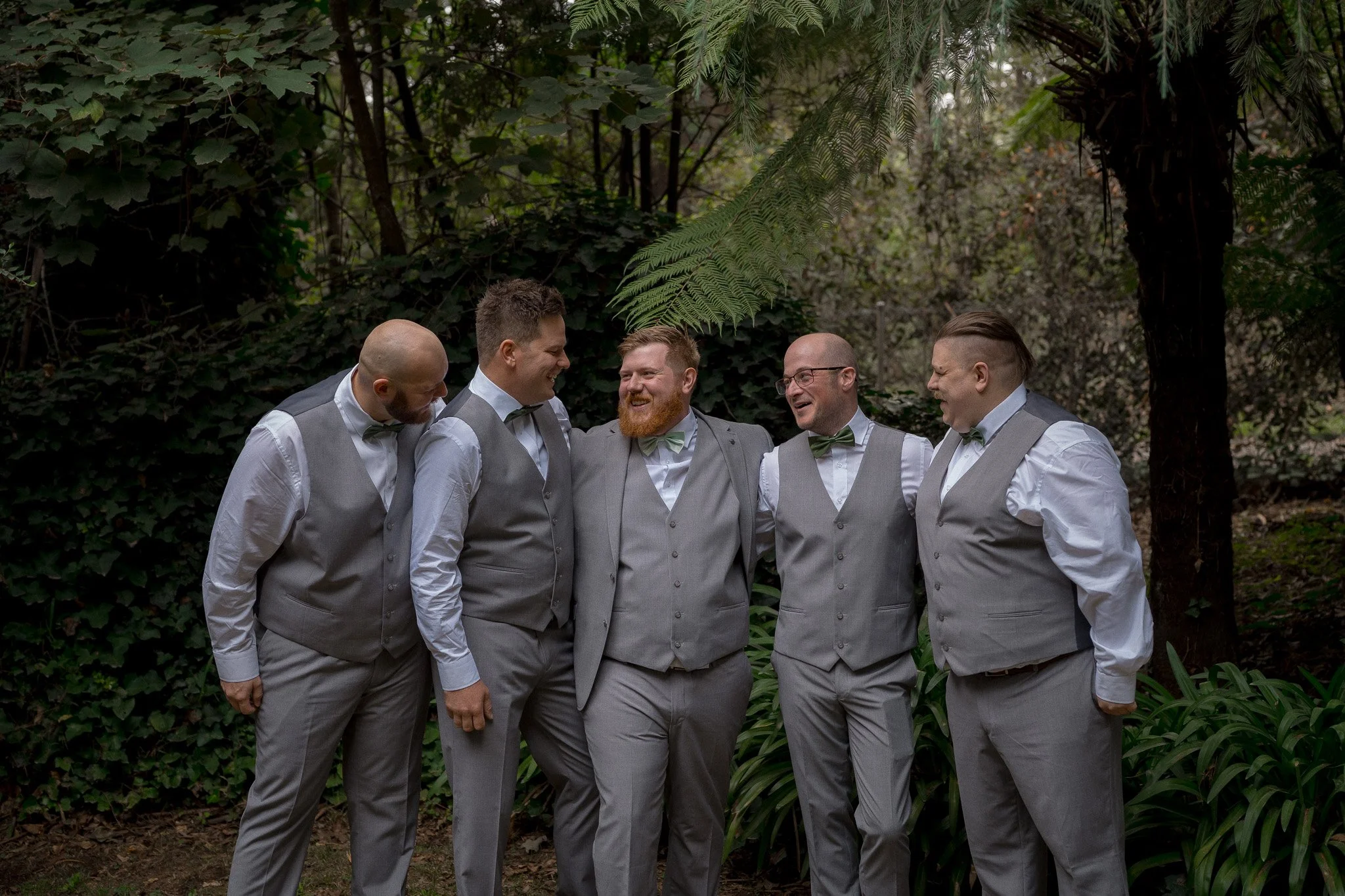 A group of six men dressed in suits and bow ties standing outdoors in front of green foliage, smiling and conversing.