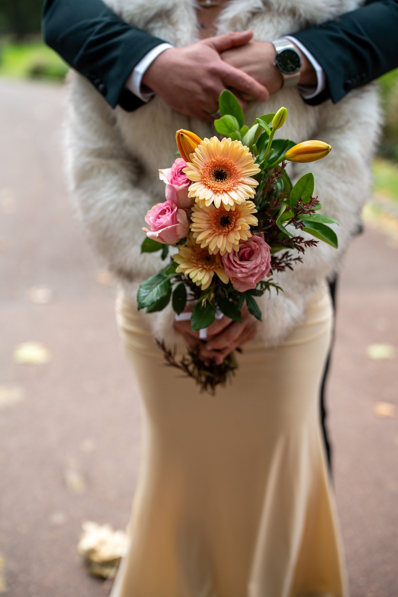 Person dressed in a beige skirt and dark jacket holding a bouquet of pink roses, orange gerbera daisies, and lilies, standing outdoors.