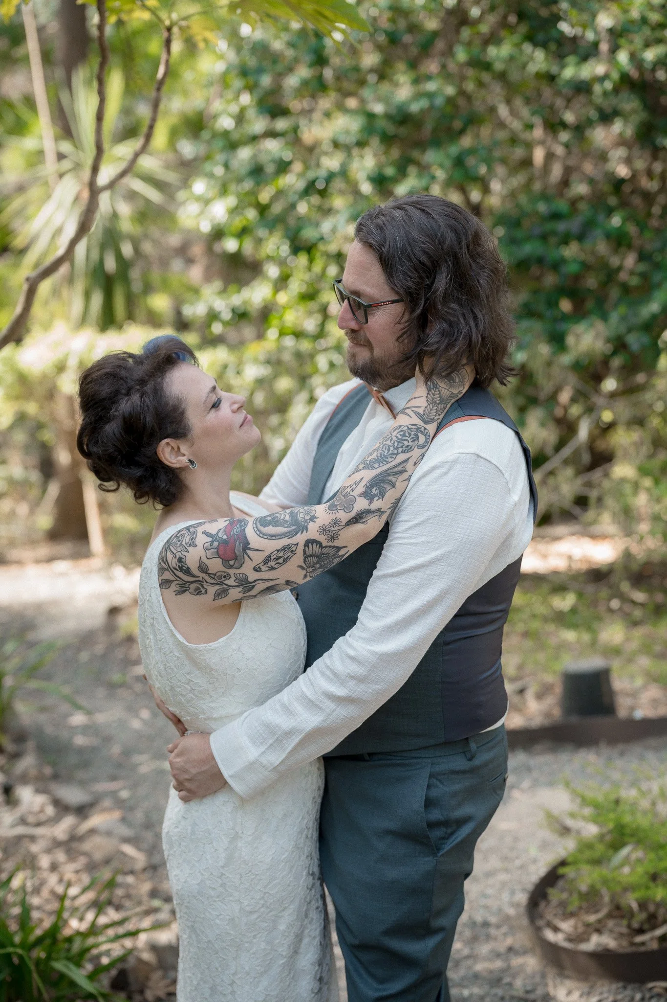 A couple, a woman with short dark hair and a man with shoulder-length dark hair and glasses, embracing outdoors surrounded by trees and plants.