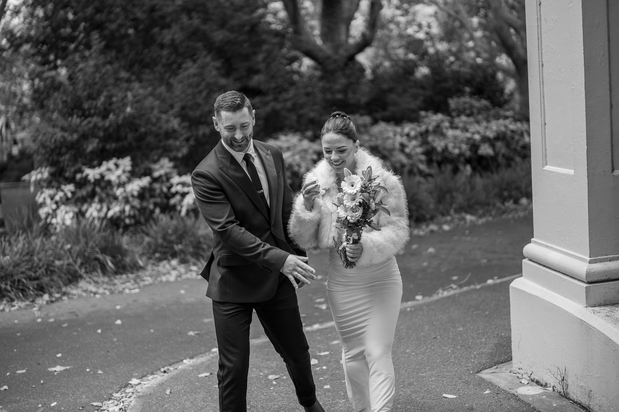 A bride and groom walking outdoors, smiling and holding hands. The bride is wearing a white dress and a fur coat, holding a bouquet of flowers. The groom is dressed in a dark suit with a tie. They are near a building with a column and there are trees and plants in the background.