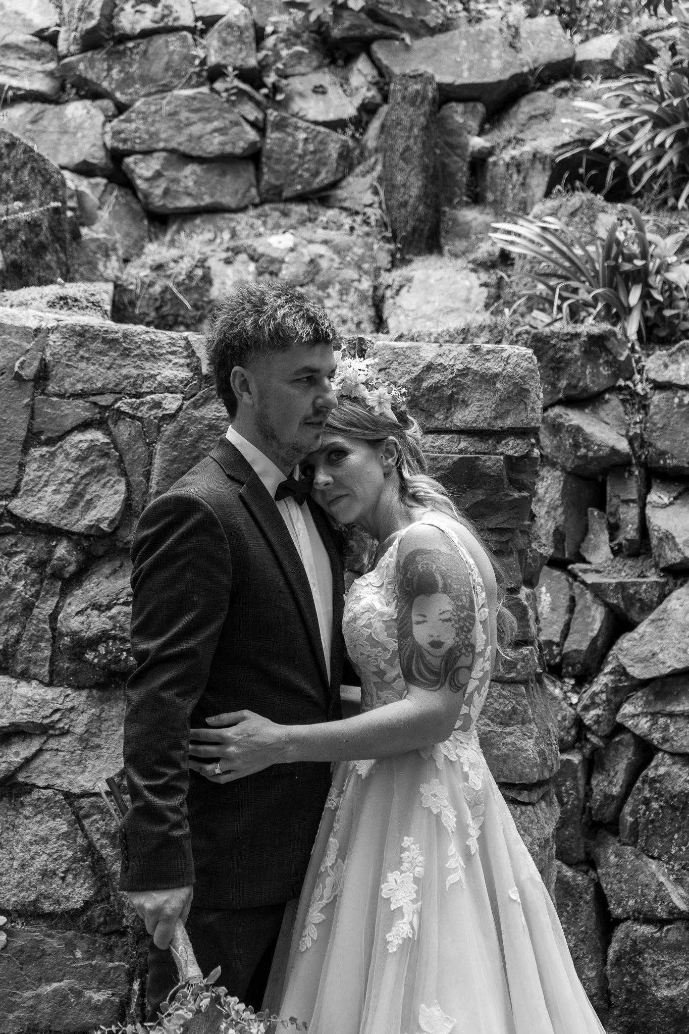A black-and-white photo of a bride and groom embracing in front of a stone wall, with the bride resting her head on the groom's shoulder and showing a tattoo of a geisha on her arm.
