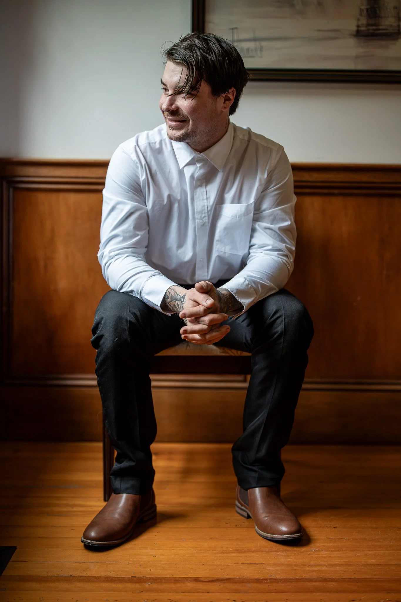 A man sitting on a wooden bench, wearing a white shirt, black pants, and brown boots, smiling and looking to his left.