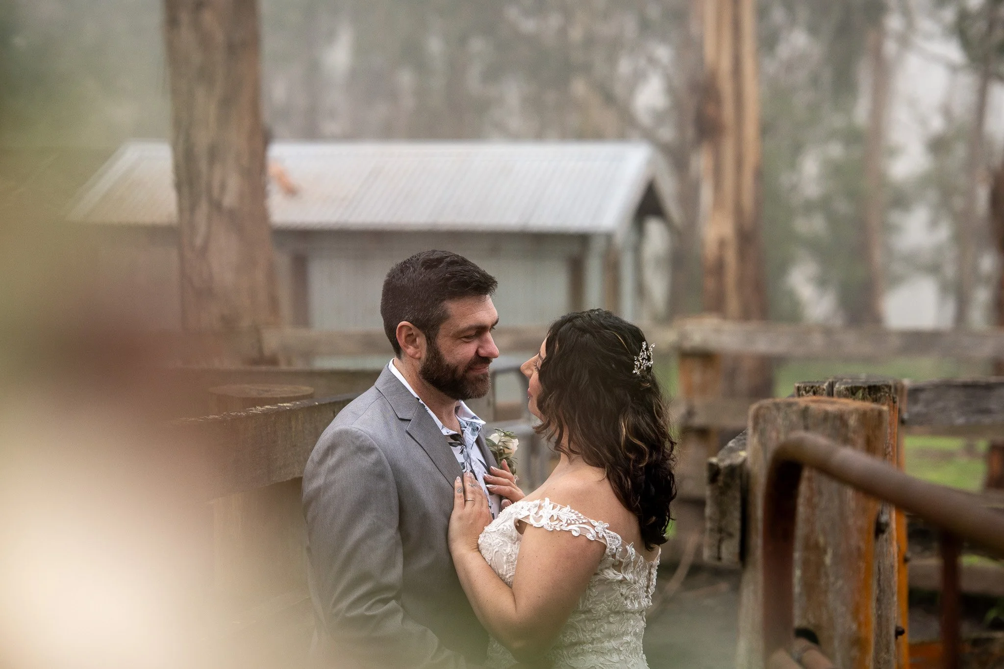 A couple dressed in wedding attire sharing an intimate moment outdoors, with trees and a rustic fence in the background.