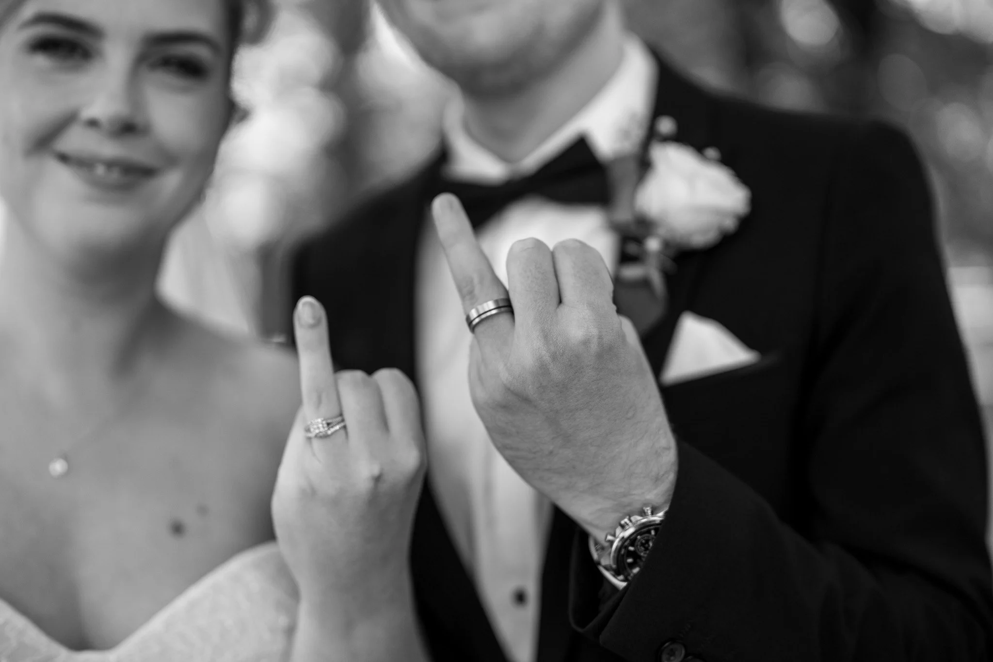 A bride and groom showing their wedding rings on their left hands, with their middle fingers raised to display the rings, smiling at the camera in black and white.