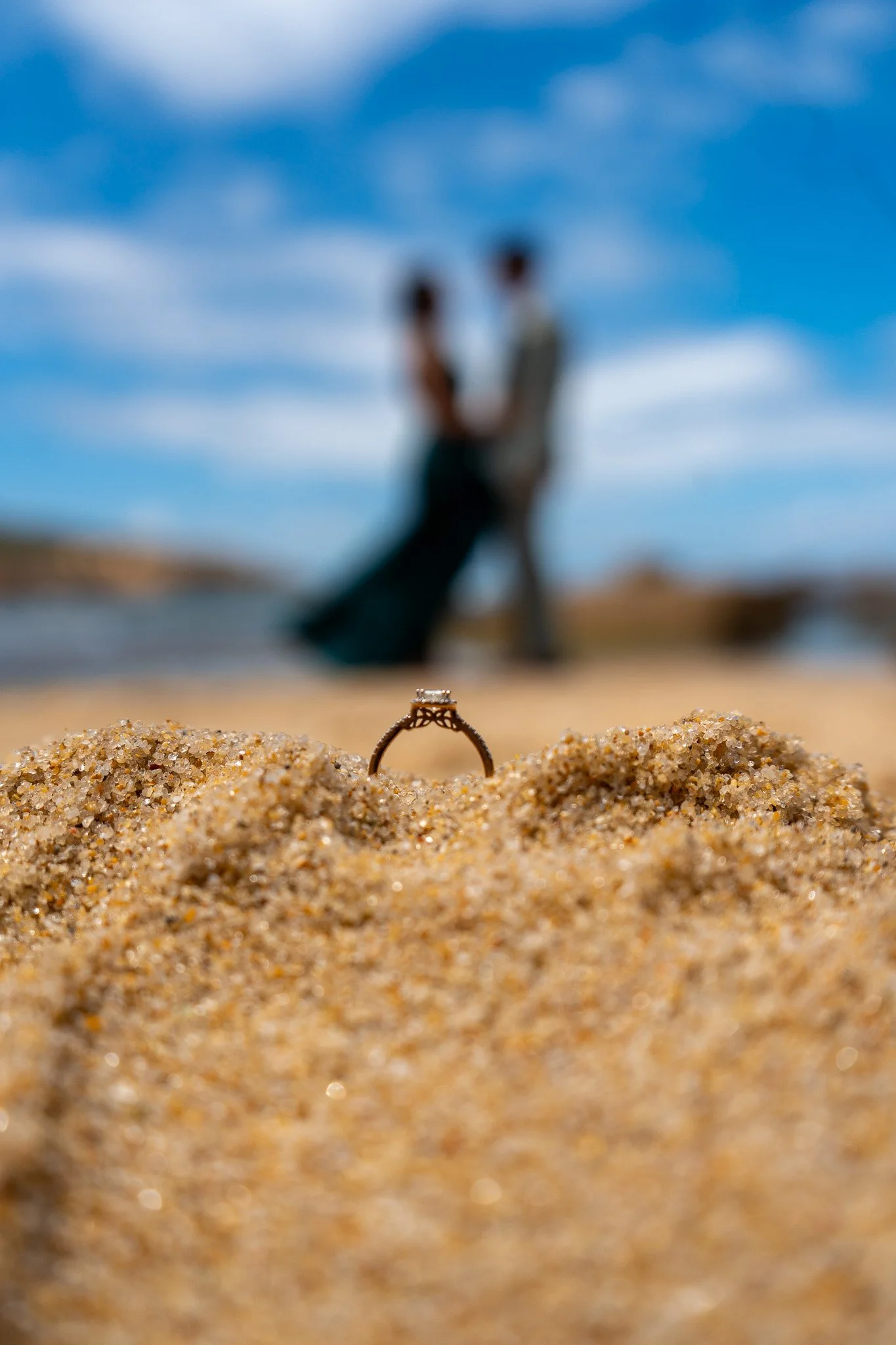 A wedding ring in the sand with a blurred couple holding hands in the background on a beach under a blue sky.