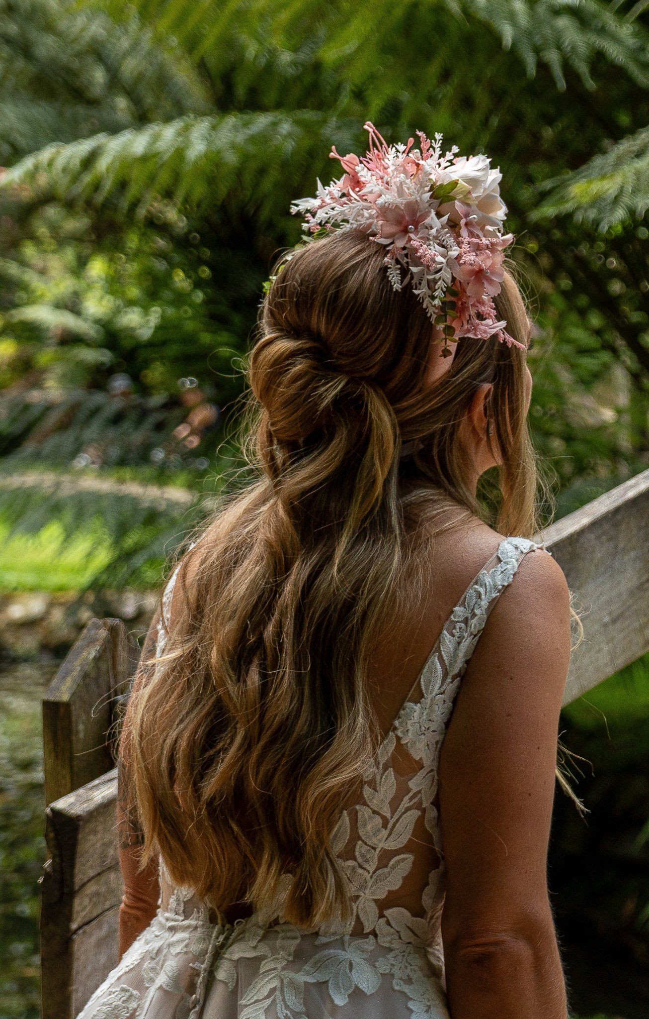 A woman with long, wavy hair styled in loose curls, wearing a lace dress with floral embroidery, is sitting on a wooden bench in a lush outdoor setting. She has a large pink flower and floral arrangement headpiece on her head.