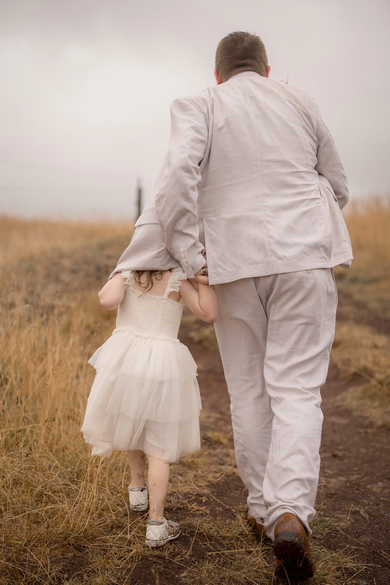 A man and young girl walking away on a dirt path through a grassy field, with the girl holding onto the man's leg.