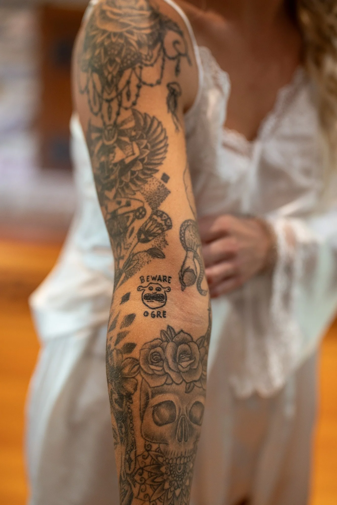 A person's arm with multiple tattoos, including a skull, roses, a woman's face, and a warning label that says 'BEWARE OGRE', with part of their torso and white clothing visible in the background.