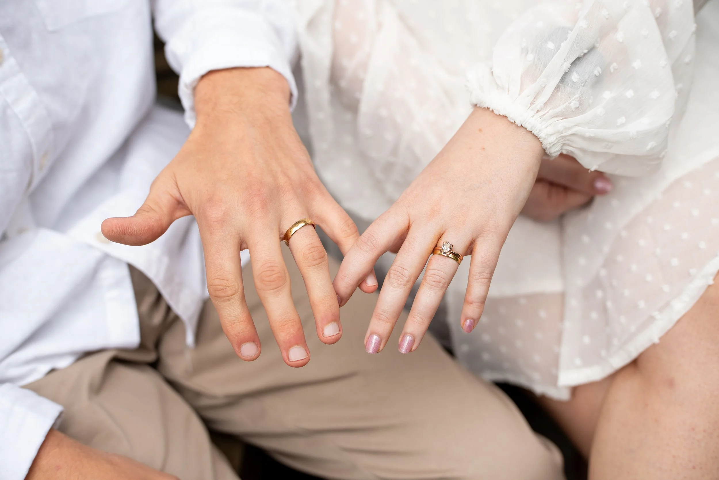 Close-up of a couple showing their hands and wedding rings. The man wears a plain gold band, and the woman has a diamond engagement ring and a gold wedding band.