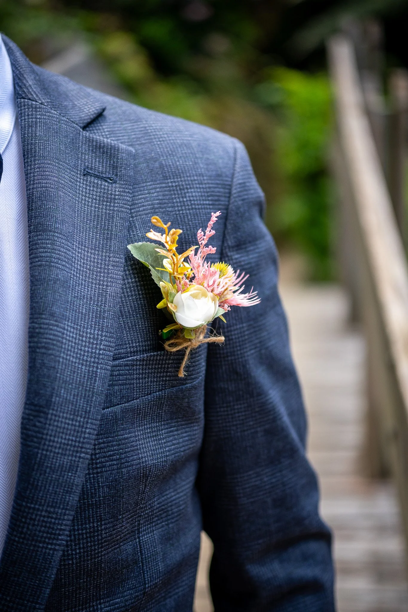 Close-up of a person wearing a blue checked suit with a floral boutonniere on the lapel, standing outdoors near a wooden bridge with greenery in the background.