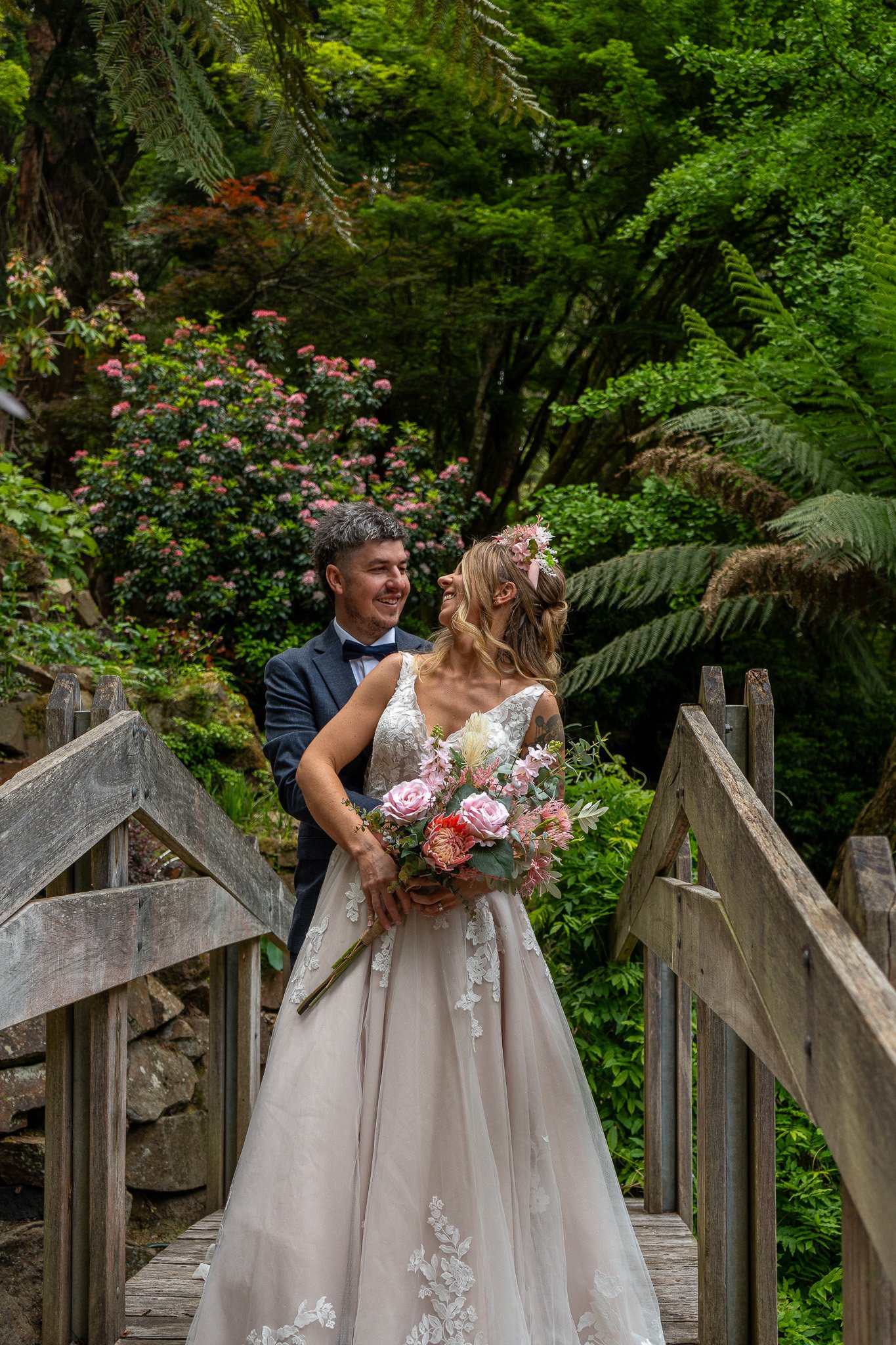A bride and groom standing on a wooden bridge surrounded by lush green foliage, with the bride holding a bouquet of pink and white flowers.