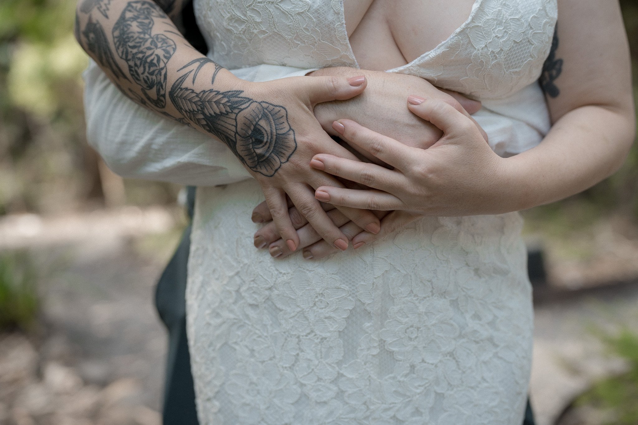 Close-up of a person holding their pregnant belly with both hands, one person with tattoos on their arm, wearing a lace white dress, outdoors.