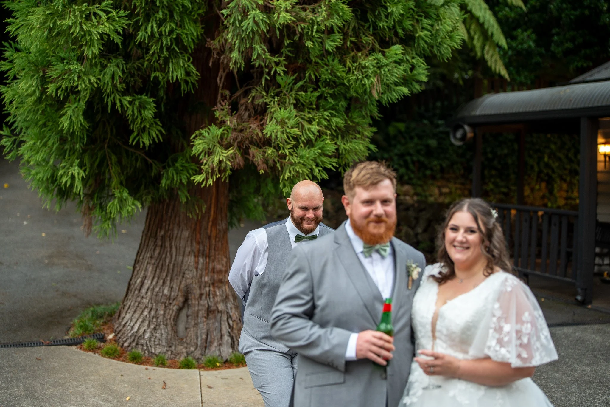 A wedding couple and two men standing outdoors near a large tree, with the groom in a gray suit holding a drink, the bride in a white lace wedding dress, and the two men dressed in vests and bow ties, smiling and enjoying the moment.