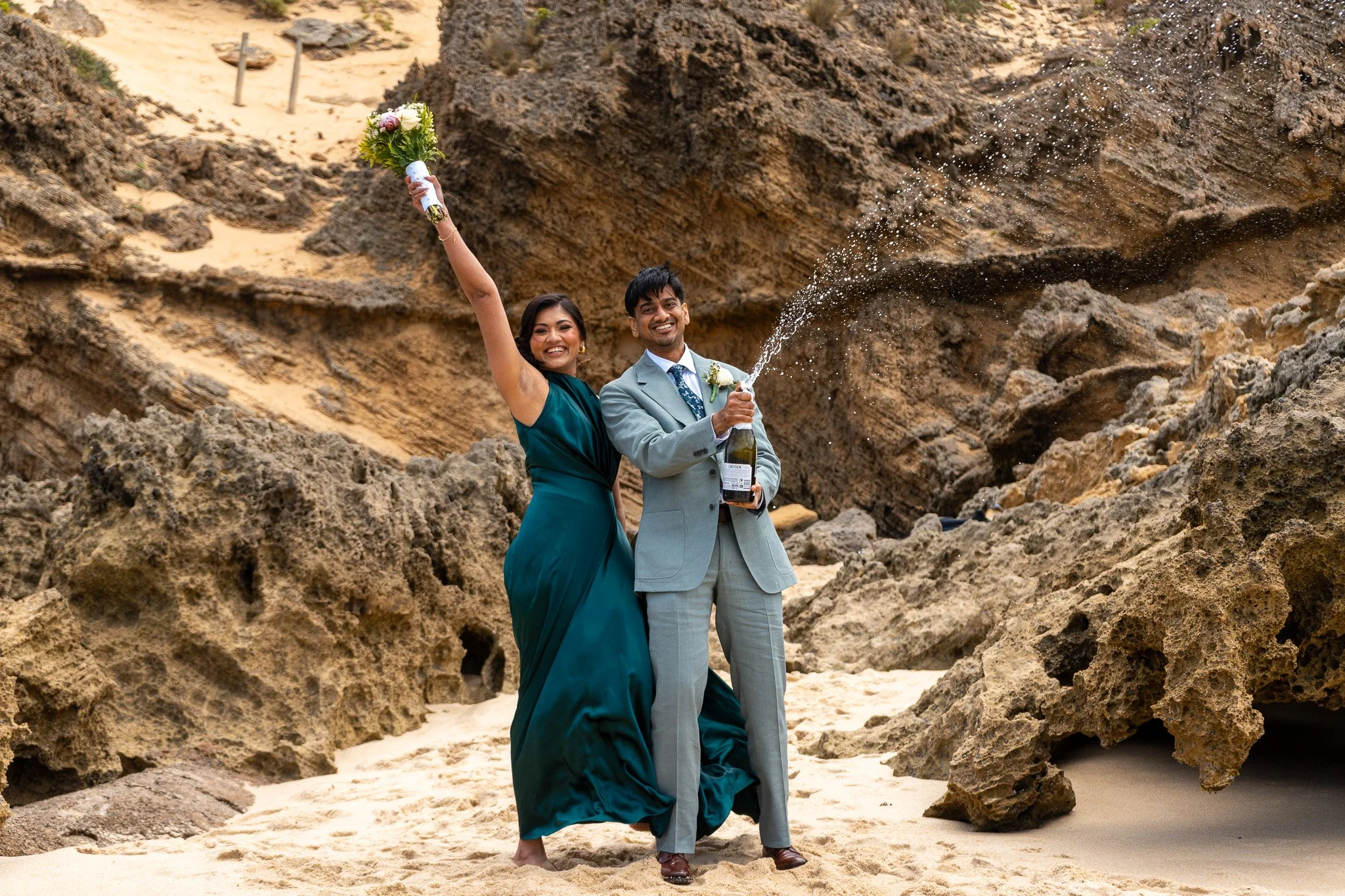 A joyful couple celebrating on a sandy beach with large rocks and cliffs in the background. The woman is wearing a teal dress and holding a bouquet in one hand and a champagne bottle in the other, while the man is dressed in a gray suit and is openin