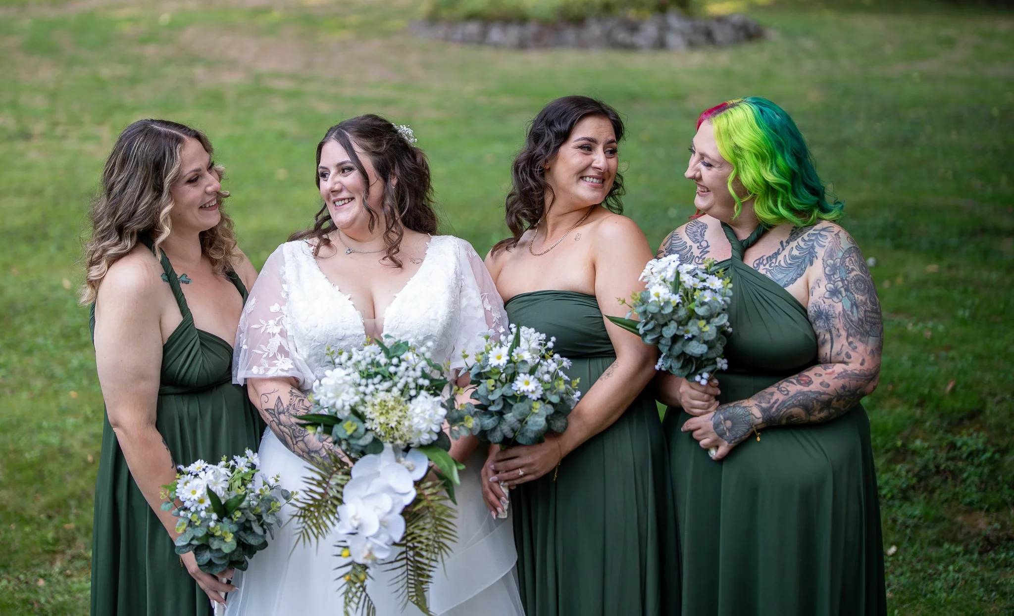 A bride and four bridesmaids standing outdoors on grass, smiling and holding bouquets of white and green flowers, with the women wearing dark green dresses and the bride in a white dress with lace details.