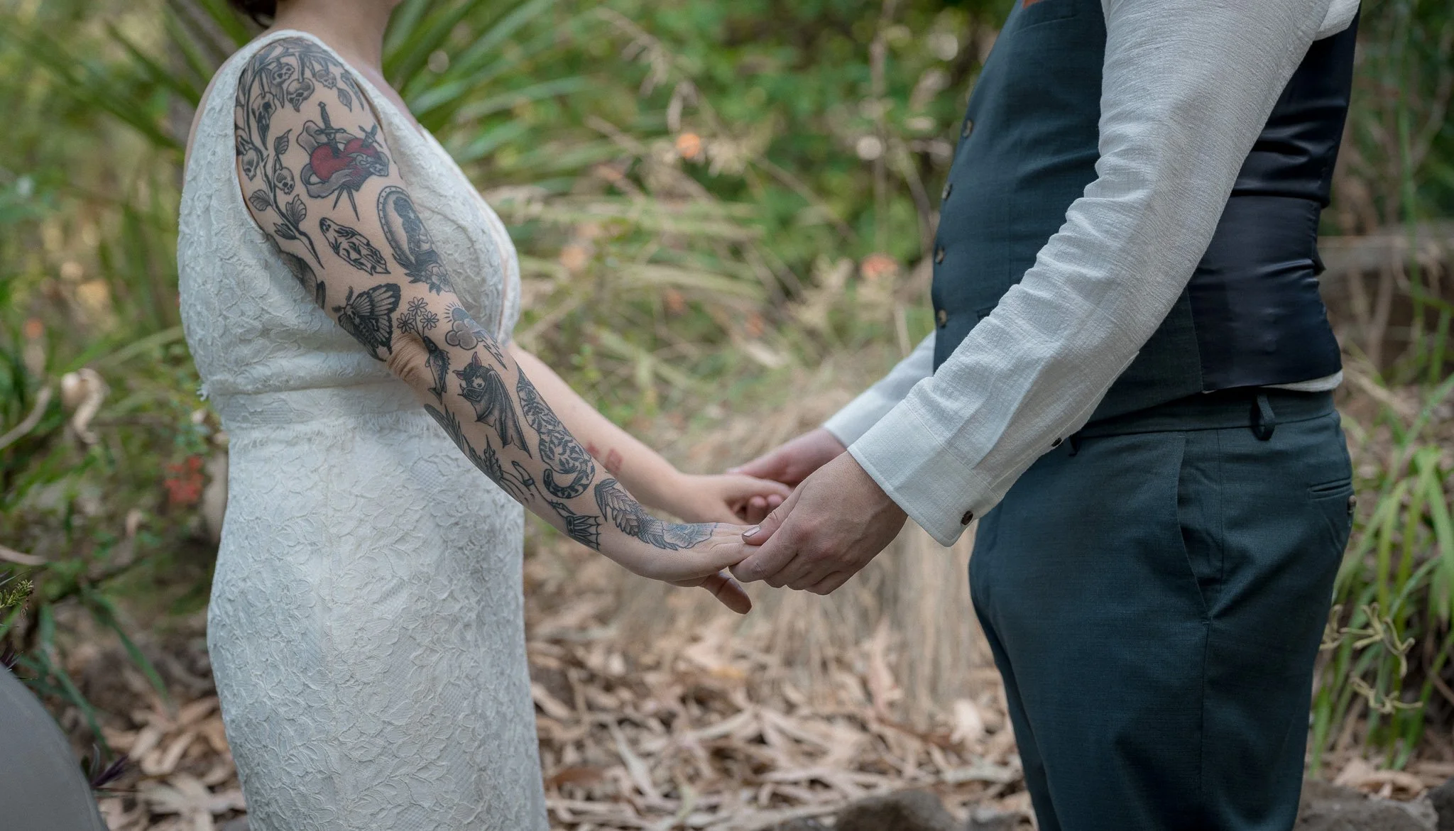 A couple holding hands during their wedding ceremony in an outdoor setting, with greenery in the background.