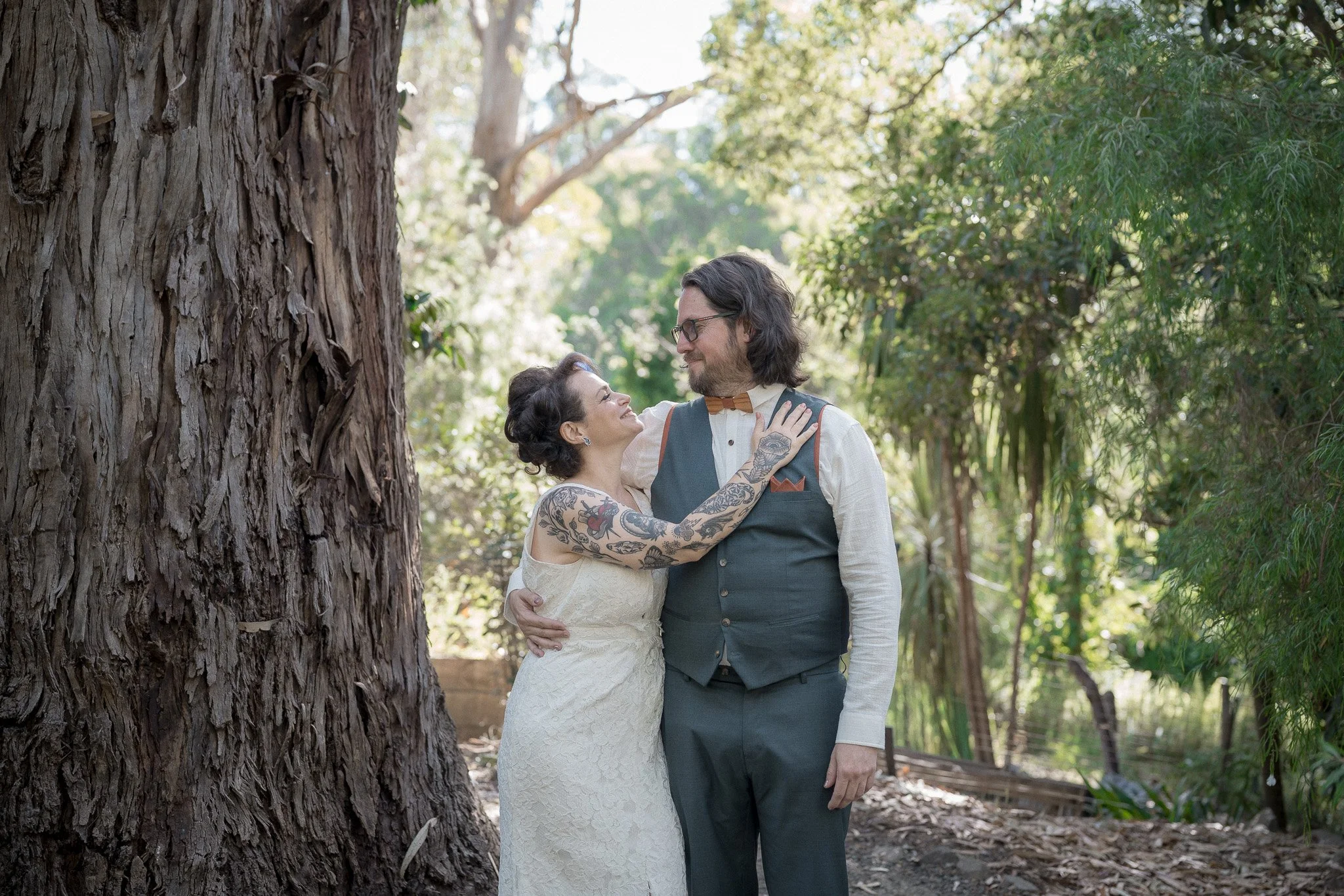A couple dressed in wedding attire standing close together outdoors among trees, sharing a loving moment.