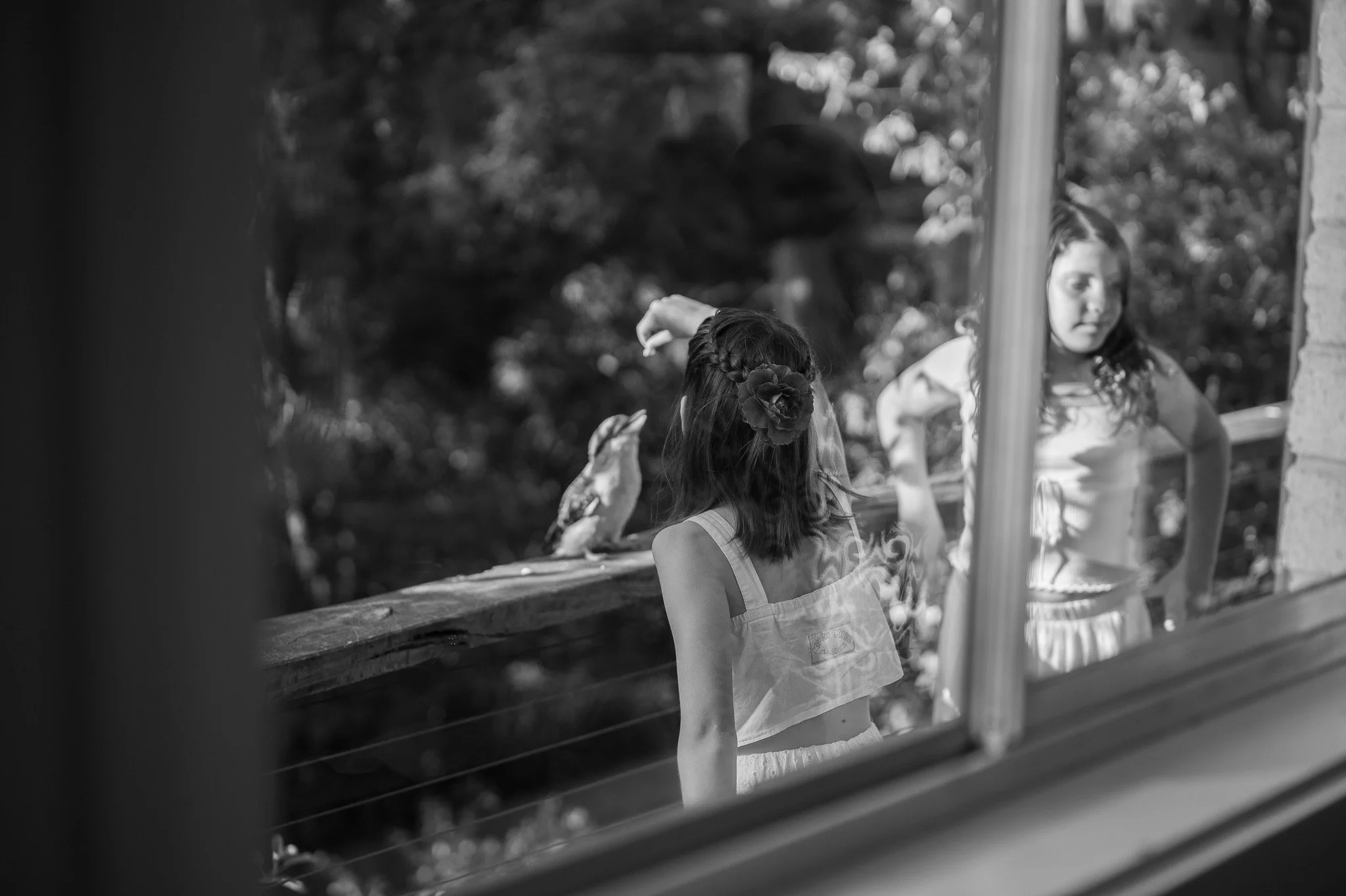 Two young girls seen through a window, one with a flower in her hair, looking at a bird on a wooden railing outside.