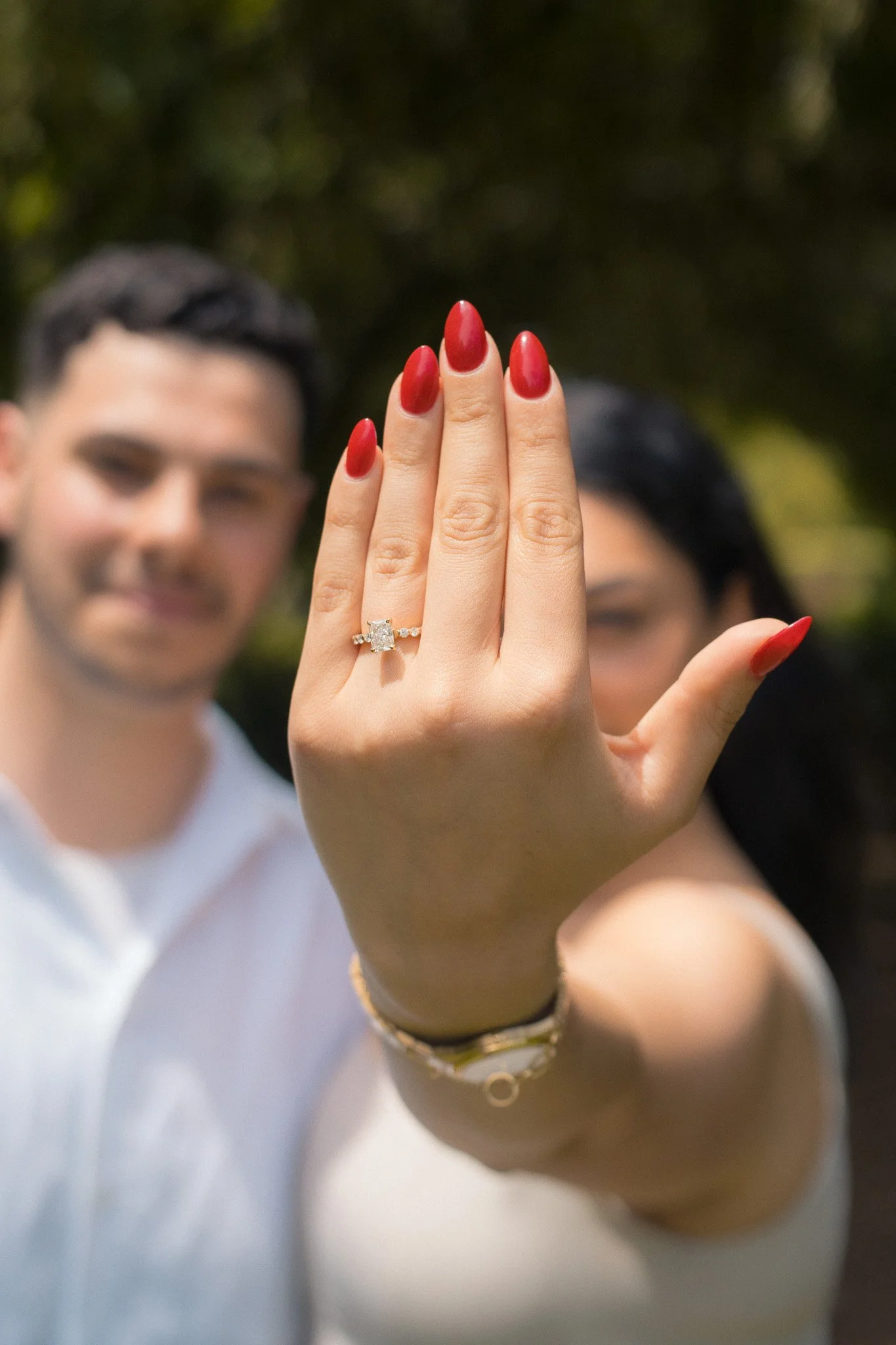 A woman showing off a diamond engagement ring on her finger with a man in the background, outdoors with trees.