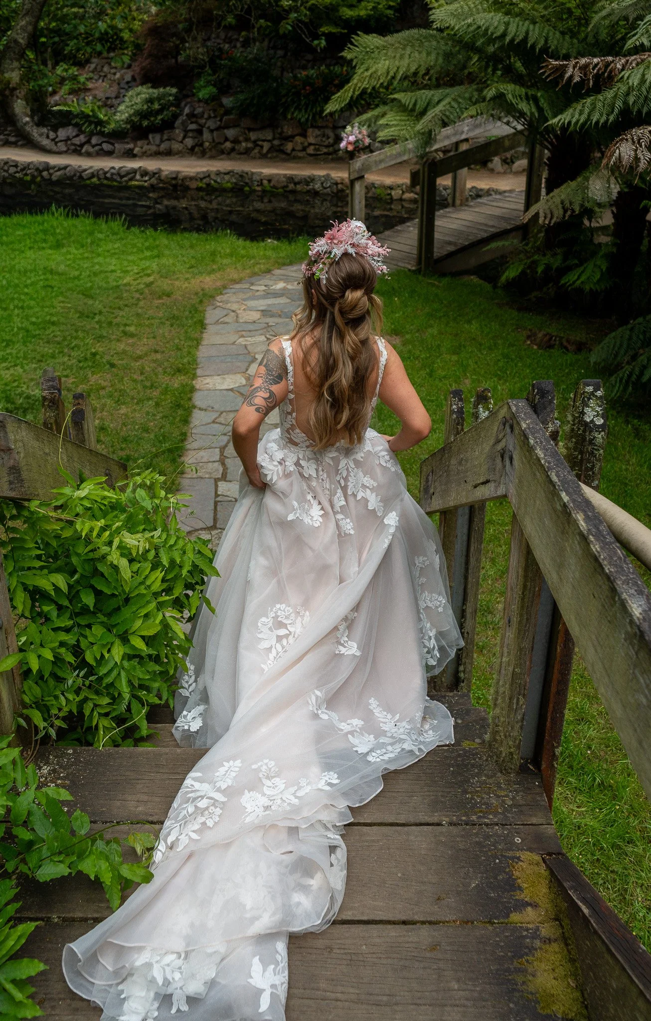 A bride in a white wedding gown with floral embroidery walking down a rustic wooden staircase in a lush green garden.