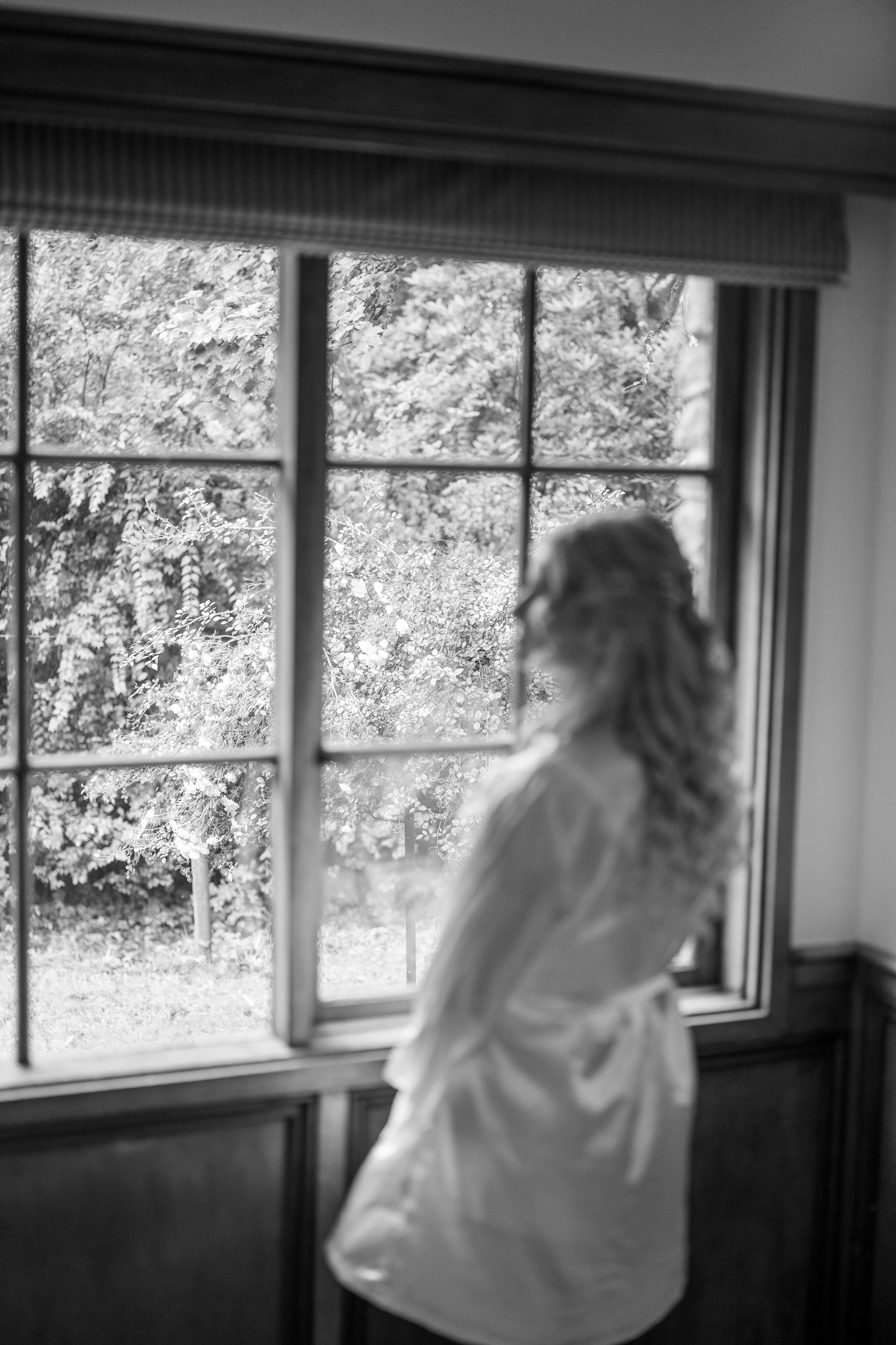 A young girl stands by a window looking outside at trees and foliage. The scene is in black and white.