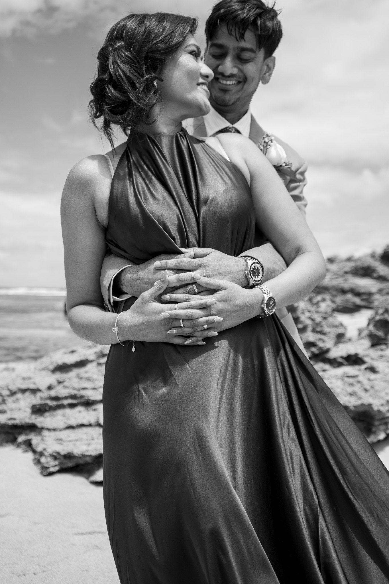 A black and white photograph of a couple embracing on a beach, with the man standing behind the woman, both smiling and looking at each other.
