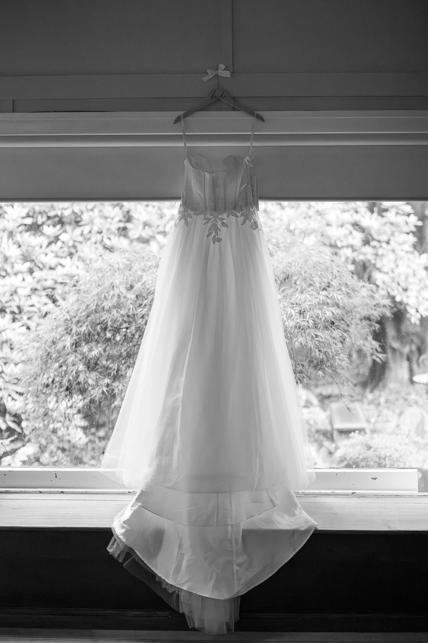 A wedding dress hanging on a hanger labeled 'bride' in front of a large window with trees outside.