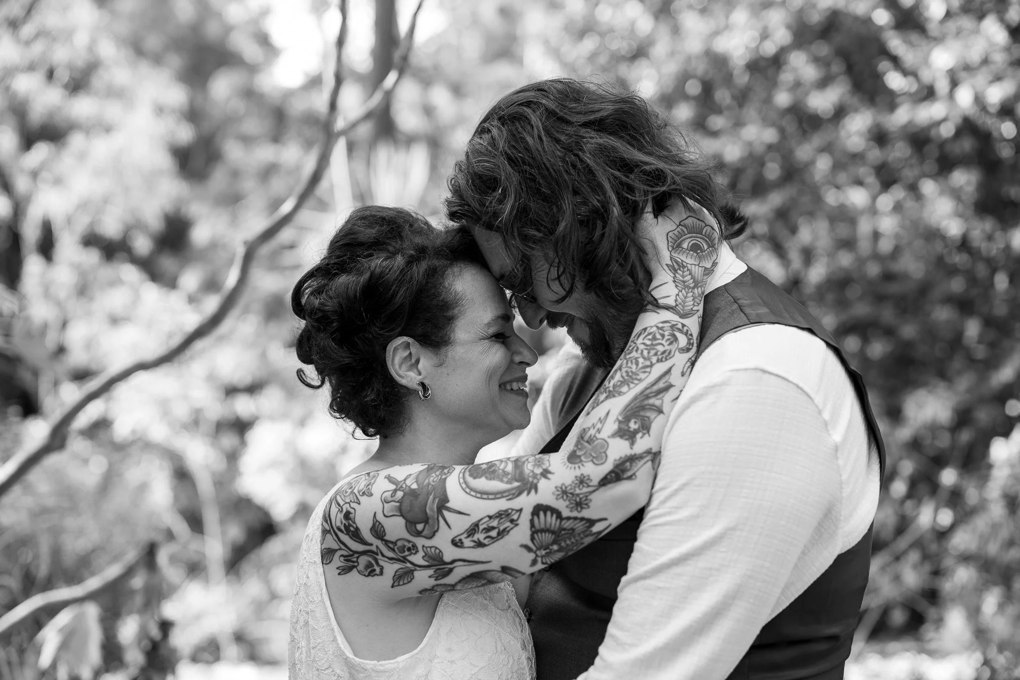 A black and white photo of a couple hugging outdoors, with their foreheads touching and smiling, surrounded by trees.