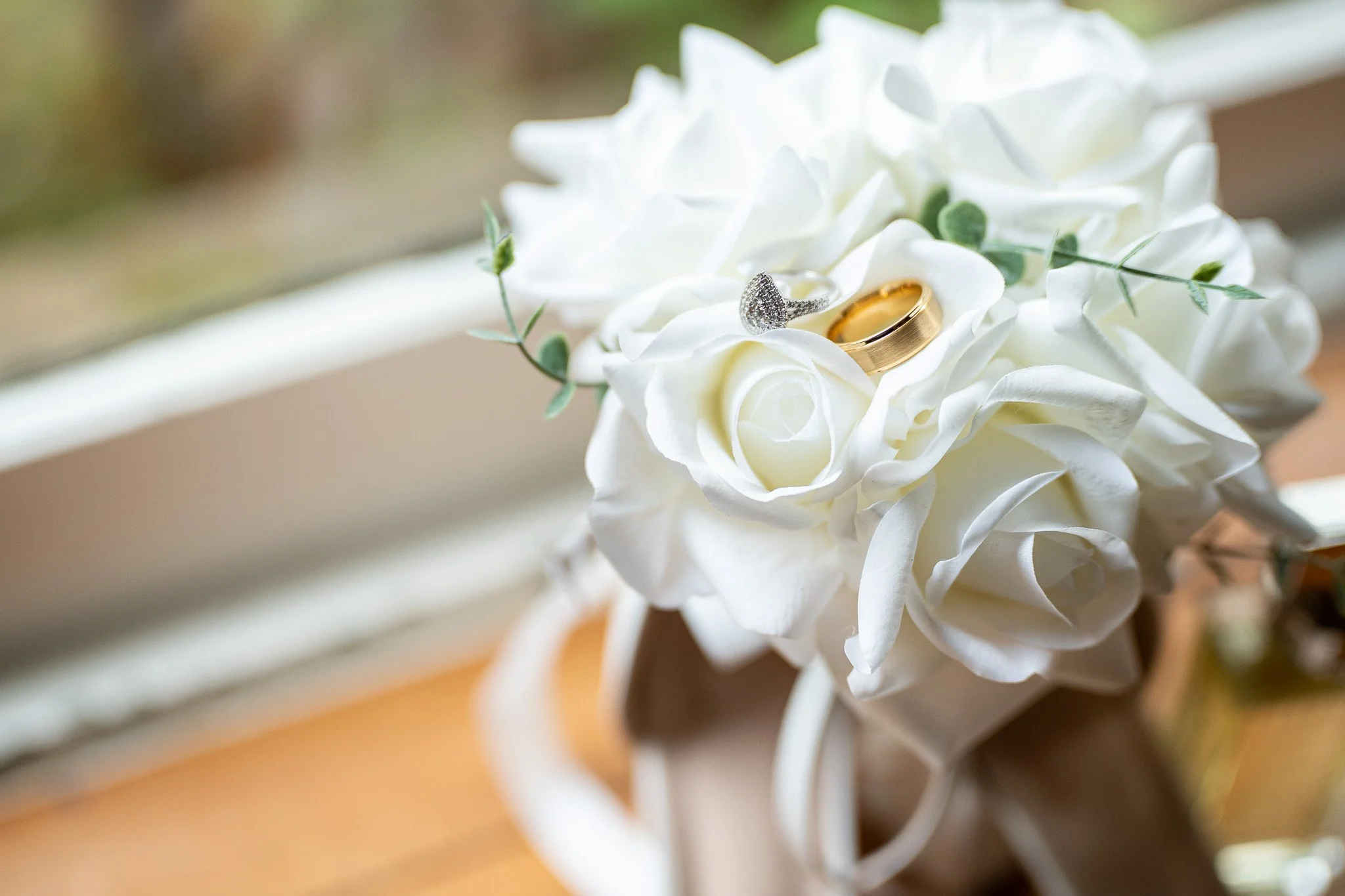 Close-up of wedding rings placed on a white flower with greenery, with a blurred background.