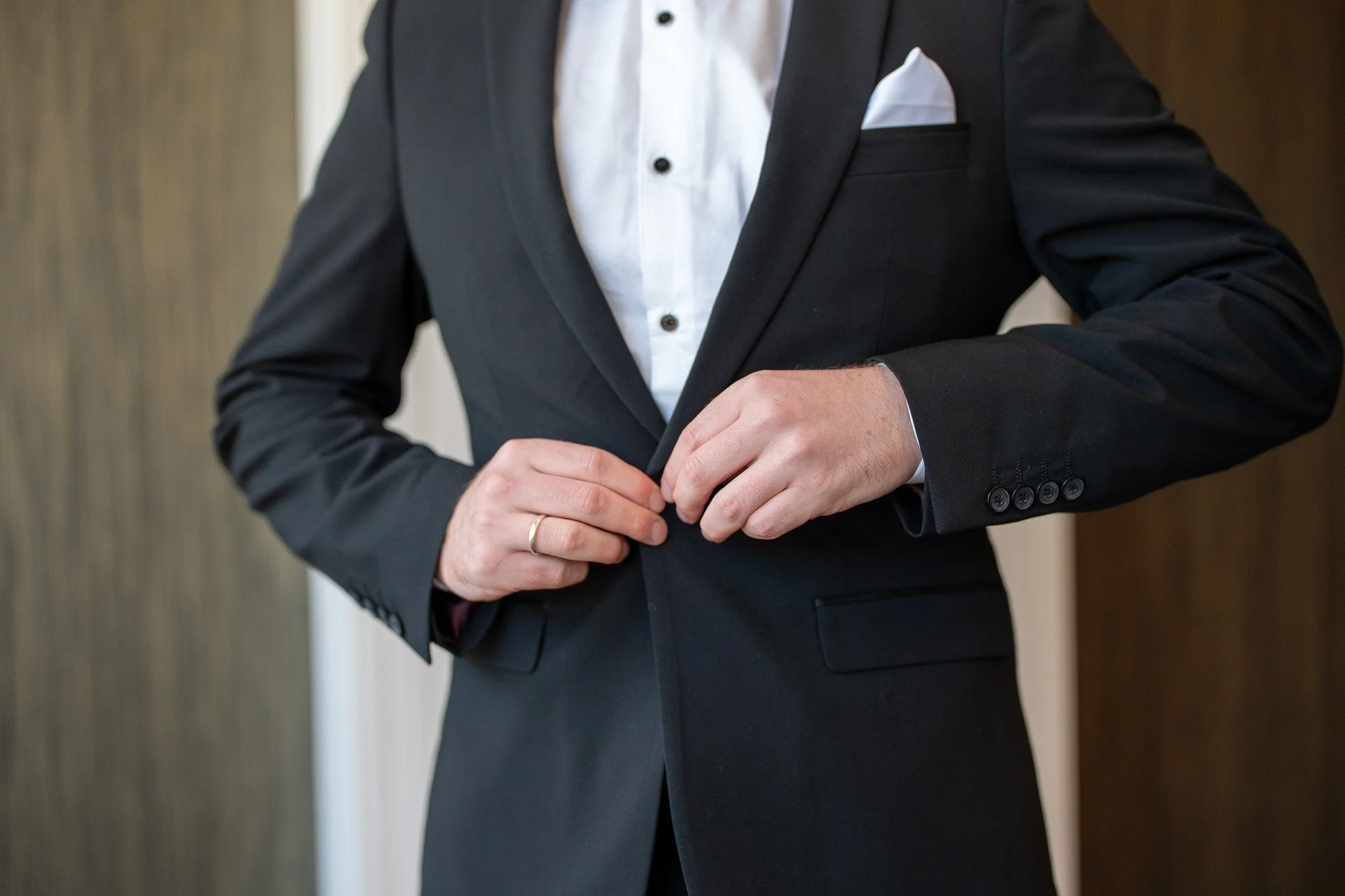 A person dressed in a black suit with a white shirt and white pocket square, adjusting their suit jacket.