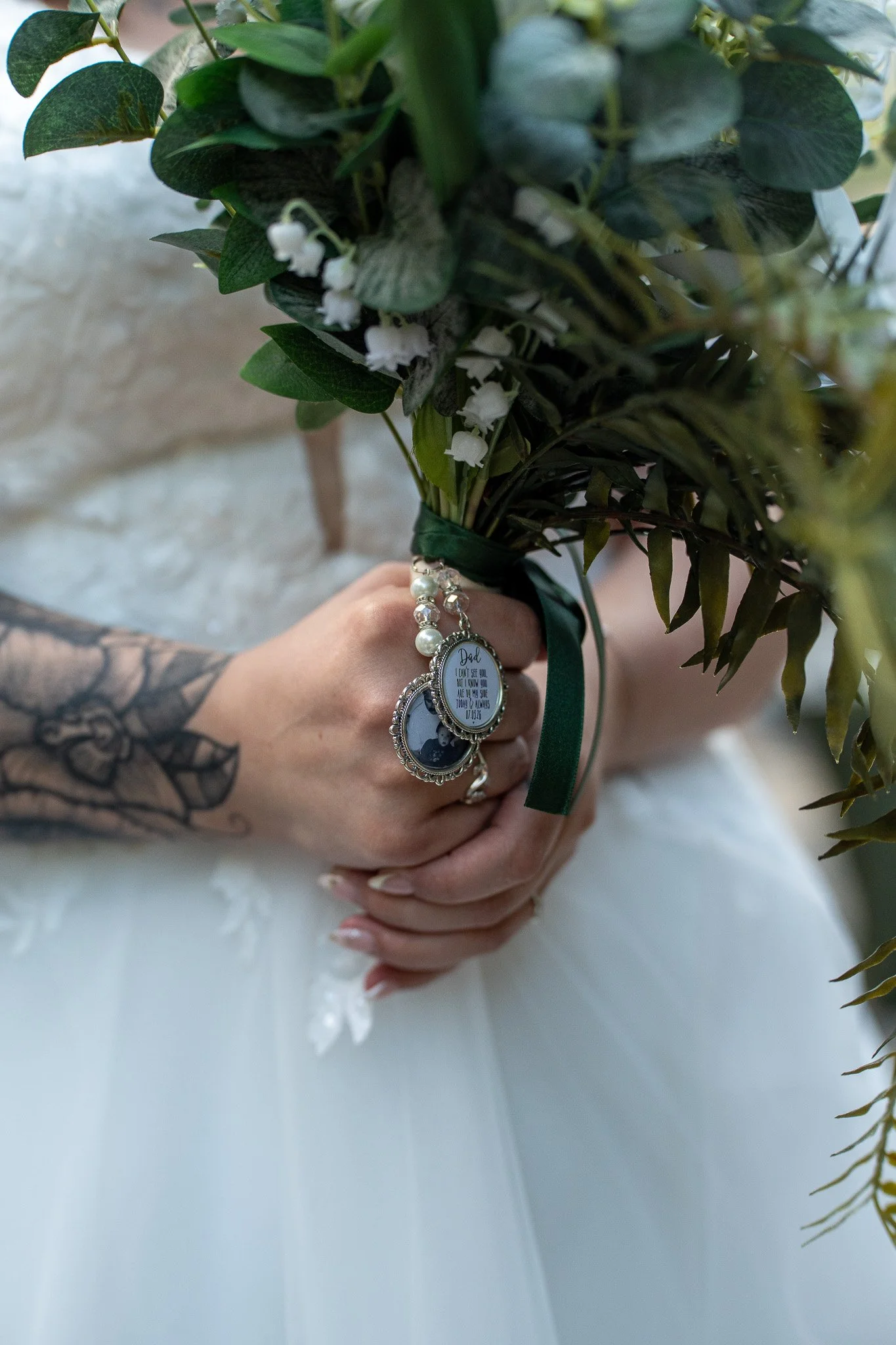 A person's hand holding a bouquet of green foliage and small white flowers, with a bracelet featuring a photo and a religious quote, and a wedding dress visible in the background.
