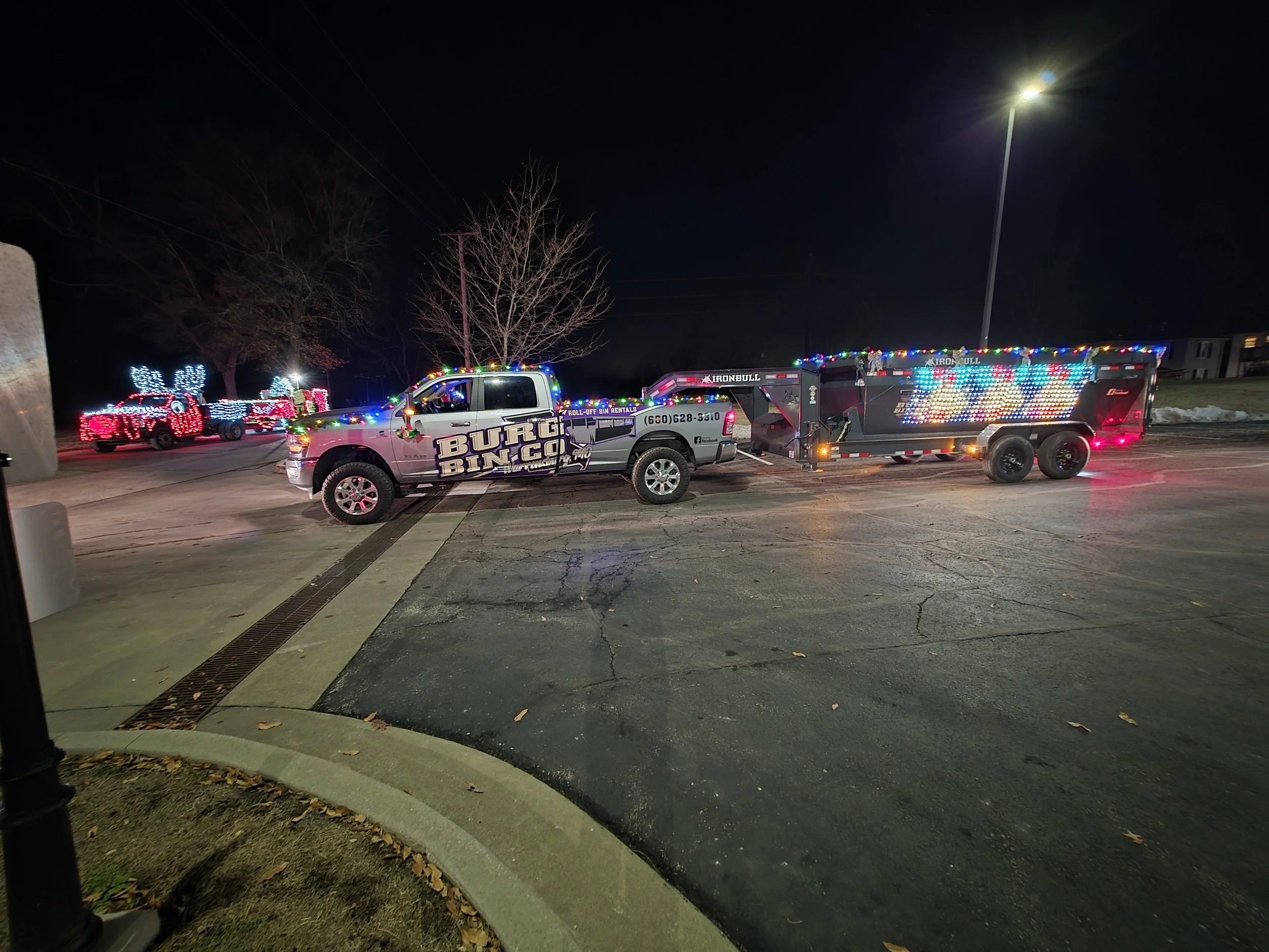 Night scene of a pickup truck decorated with colorful Christmas lights, pulling a trailer with matching lights, parked in a lot near illuminated holiday light displays of red and white reindeer and a sleigh in the background.