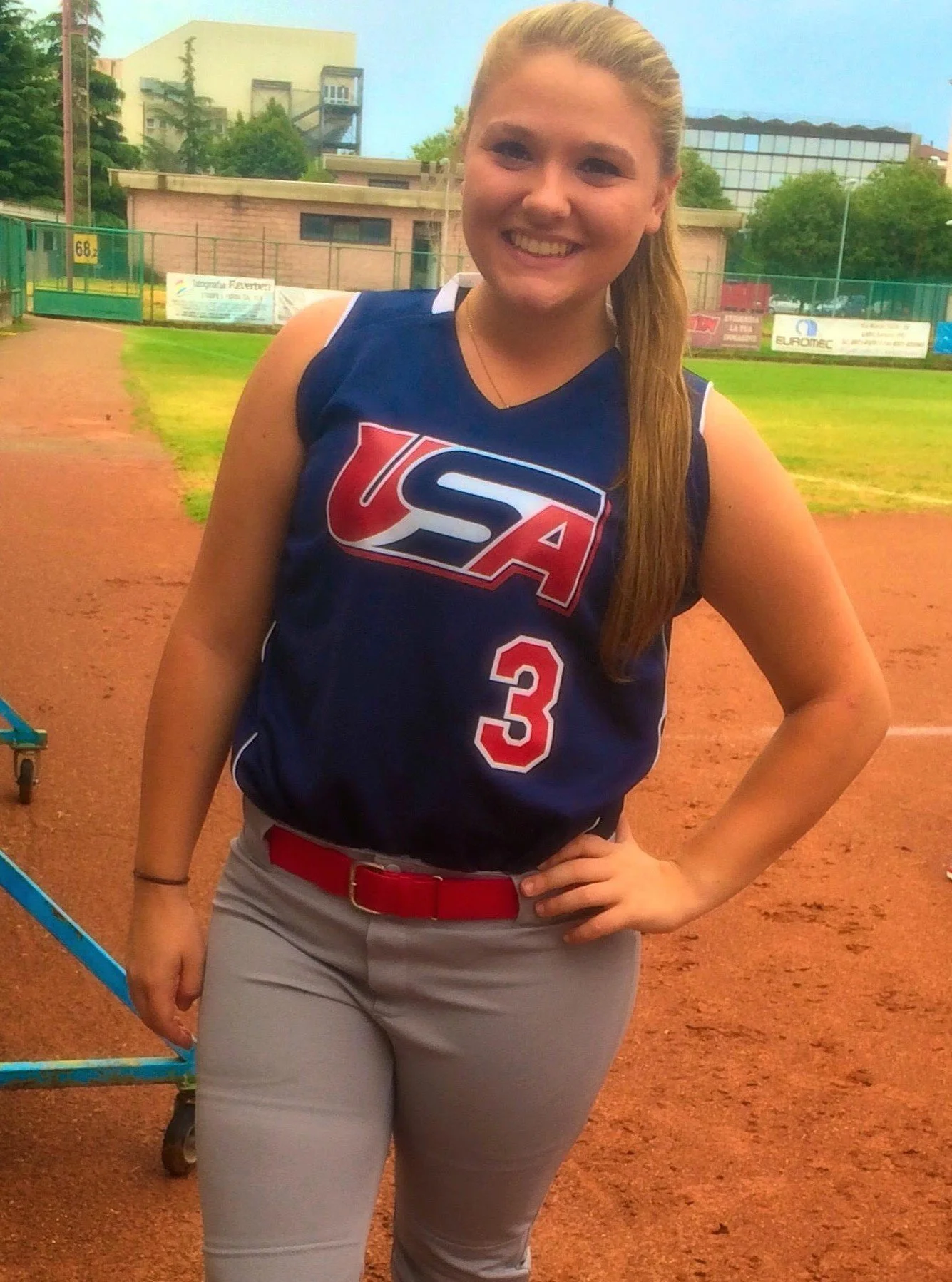 A young woman wearing a USA softball jersey with the number 3, beige pants, and a red belt, standing on a softball field with her hand on her hip and smiling.