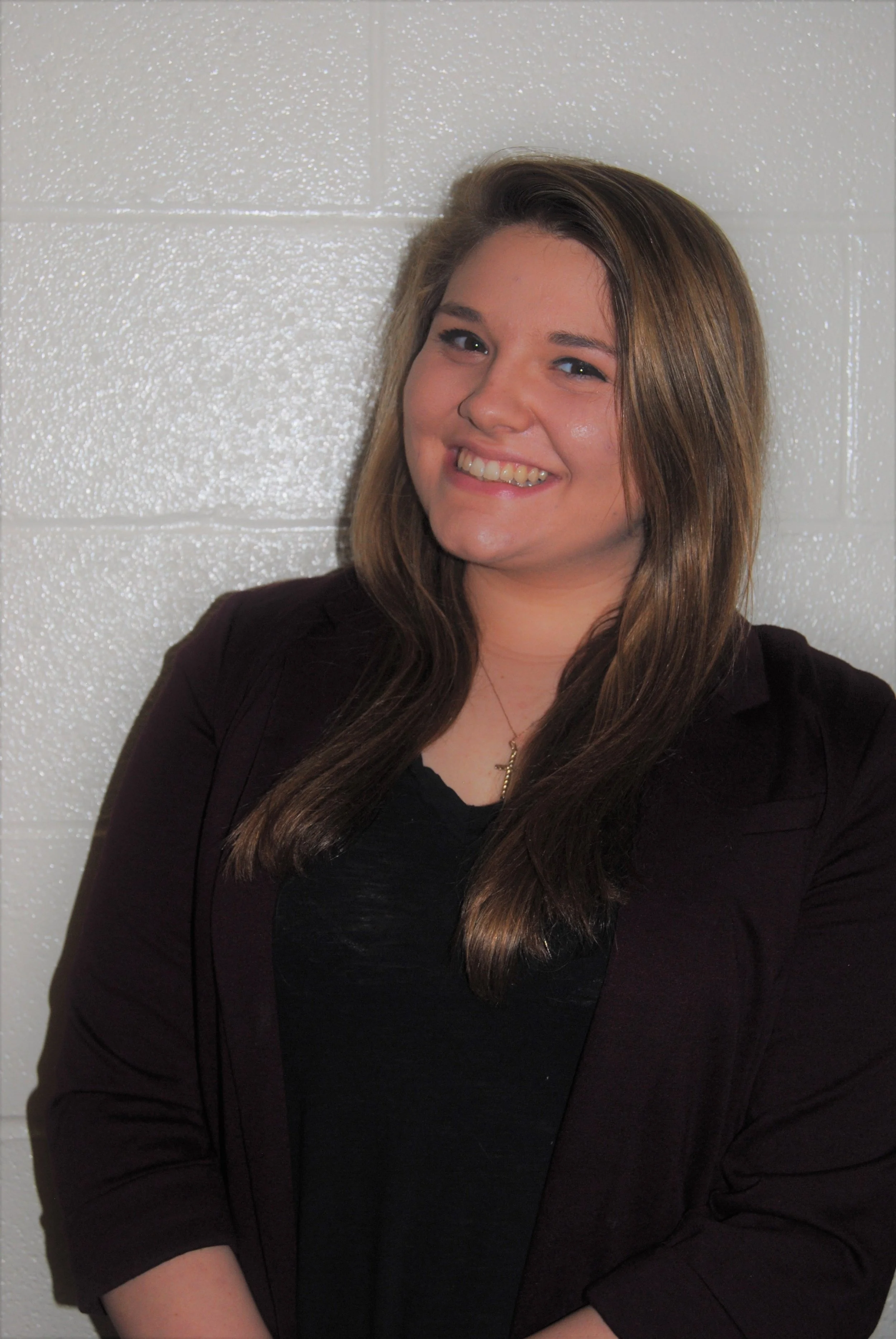 A smiling young woman with long brown hair wearing a black top and a dark blazer standing against a white textured wall.