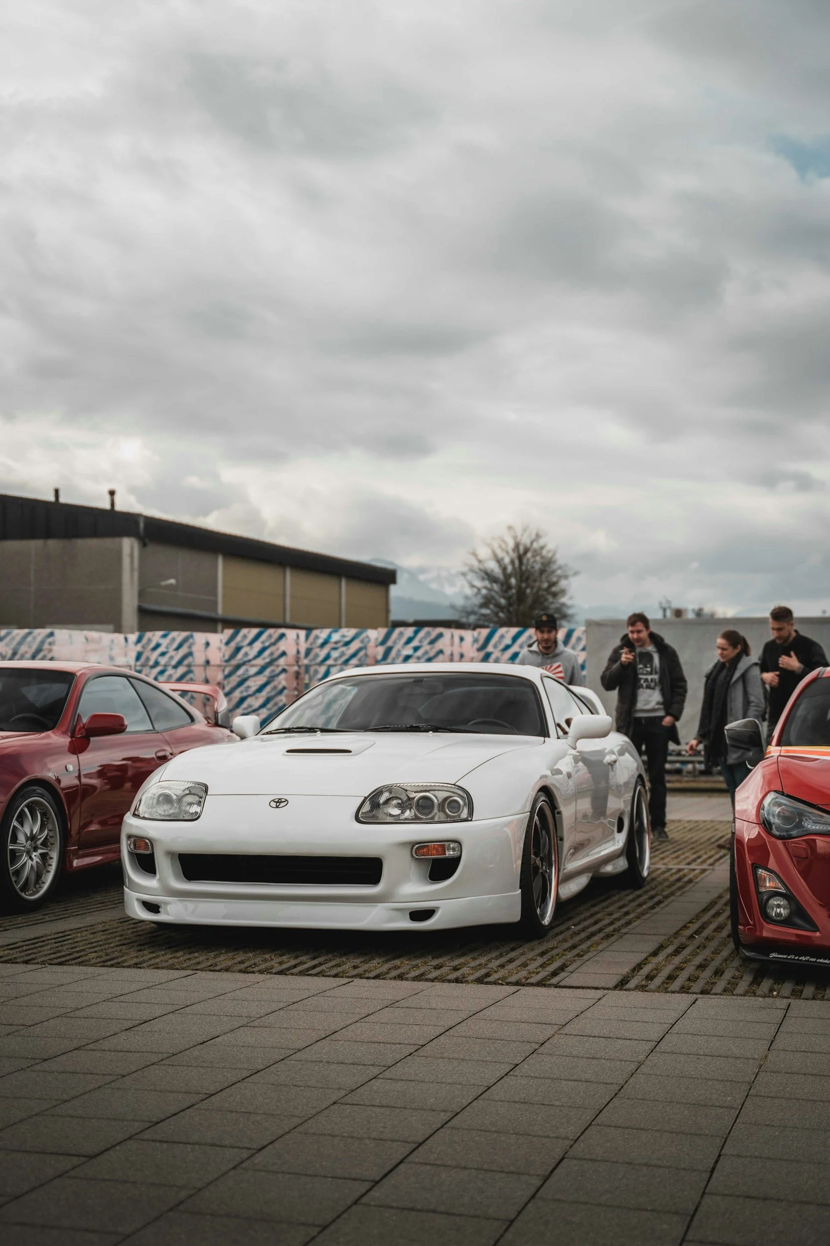 A white Toyota Supra sports car parked among other cars at a car meet, with several people standing near the vehicles, under a cloudy sky.