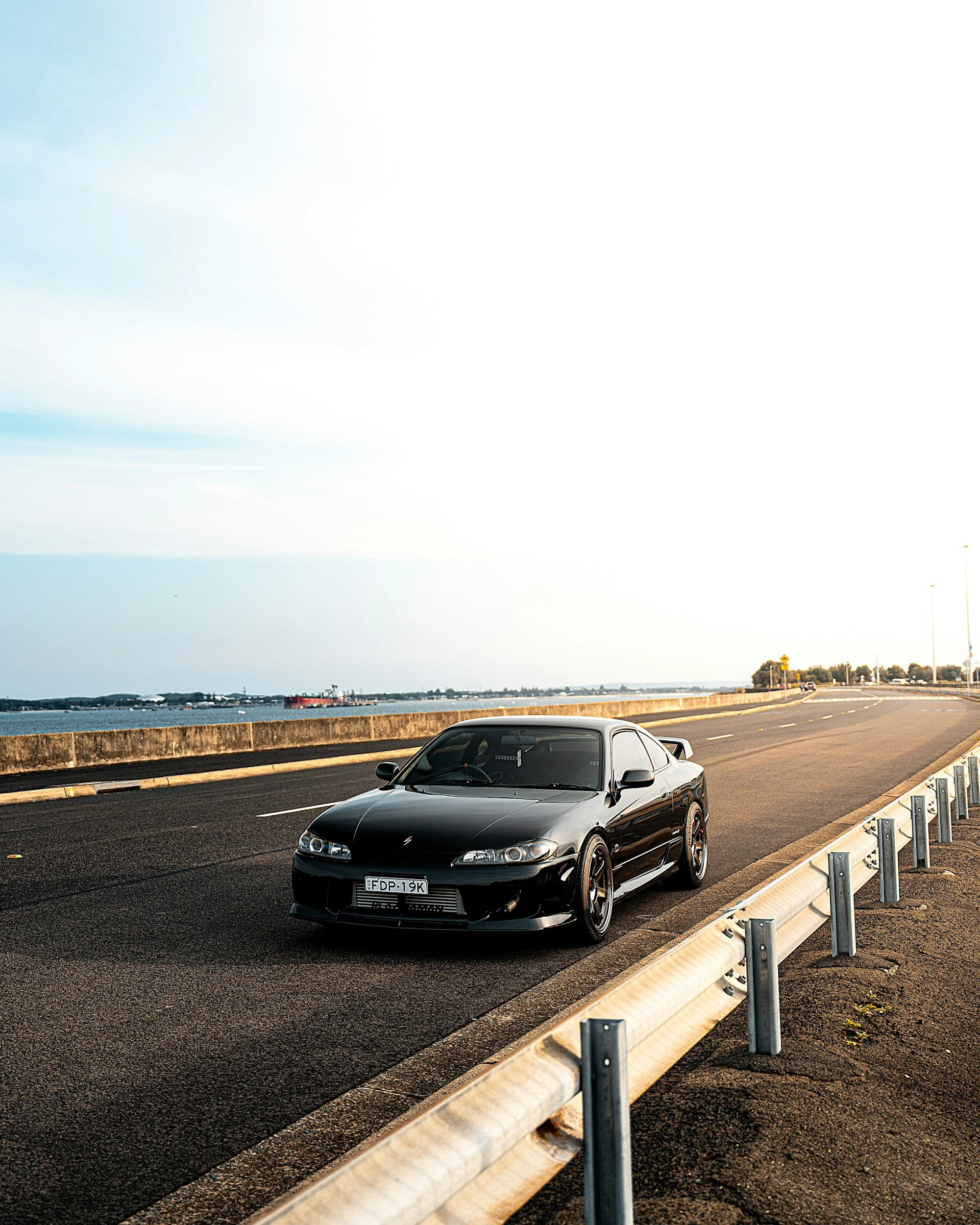 A black sports car parked on the side of an empty road by the water during sunset.