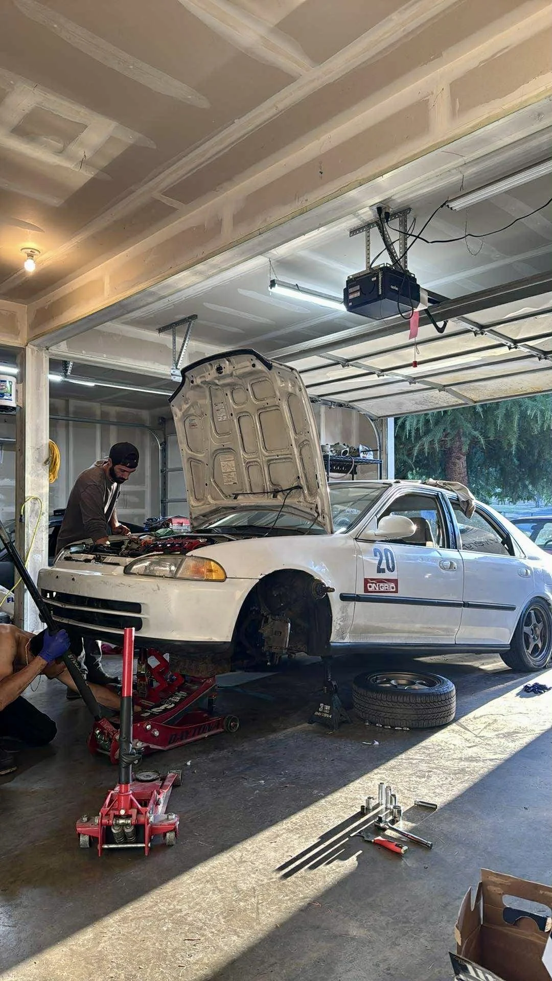 Two men working on a white car inside a garage, with the front wheel removed, the hood open, and the car raised on a jack. Tools and a spare tire are on the floor.
