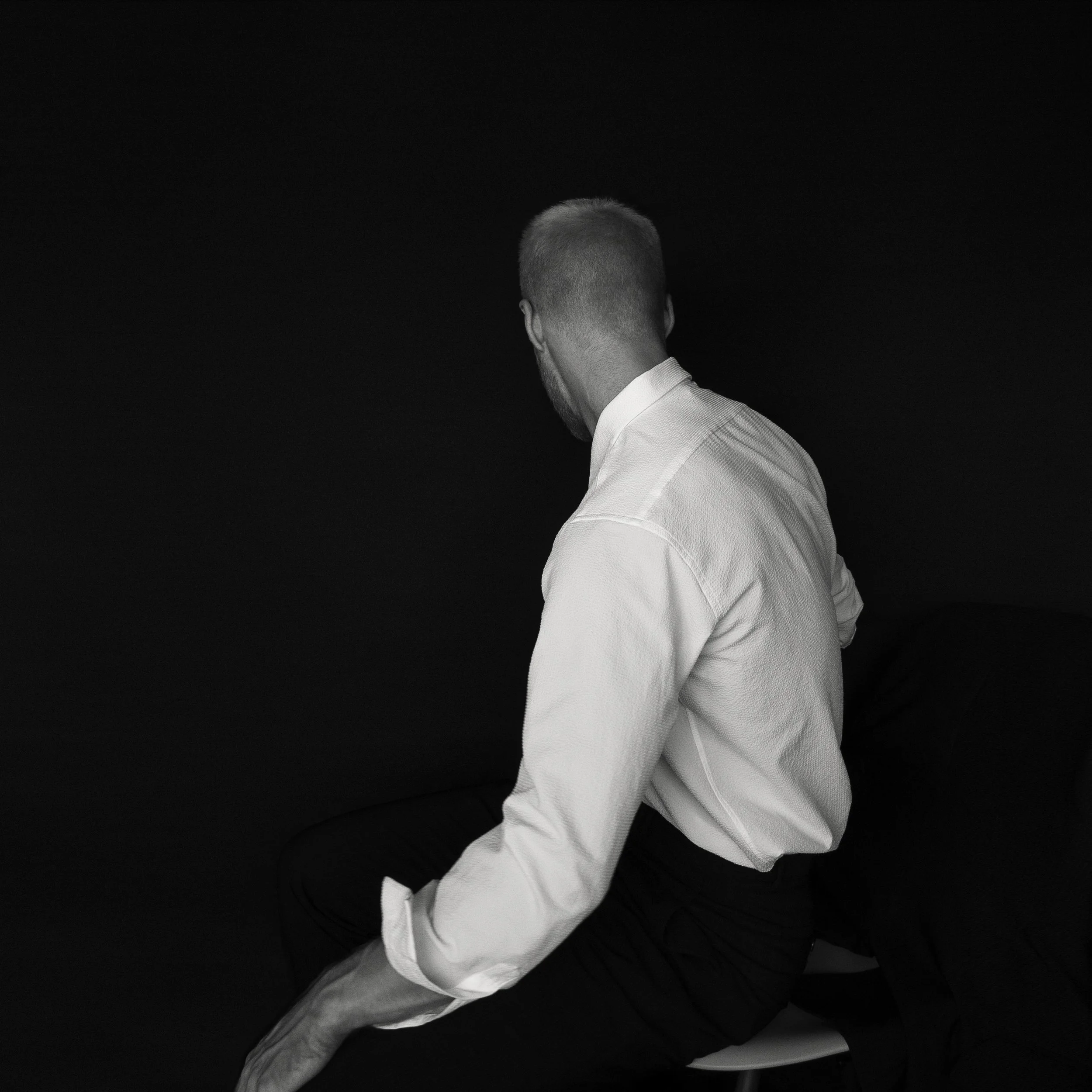 A black-and-white photo of a man with short hair, wearing a white shirt, sitting on a stool against a black background. The man is facing away from the camera and looking to the left.