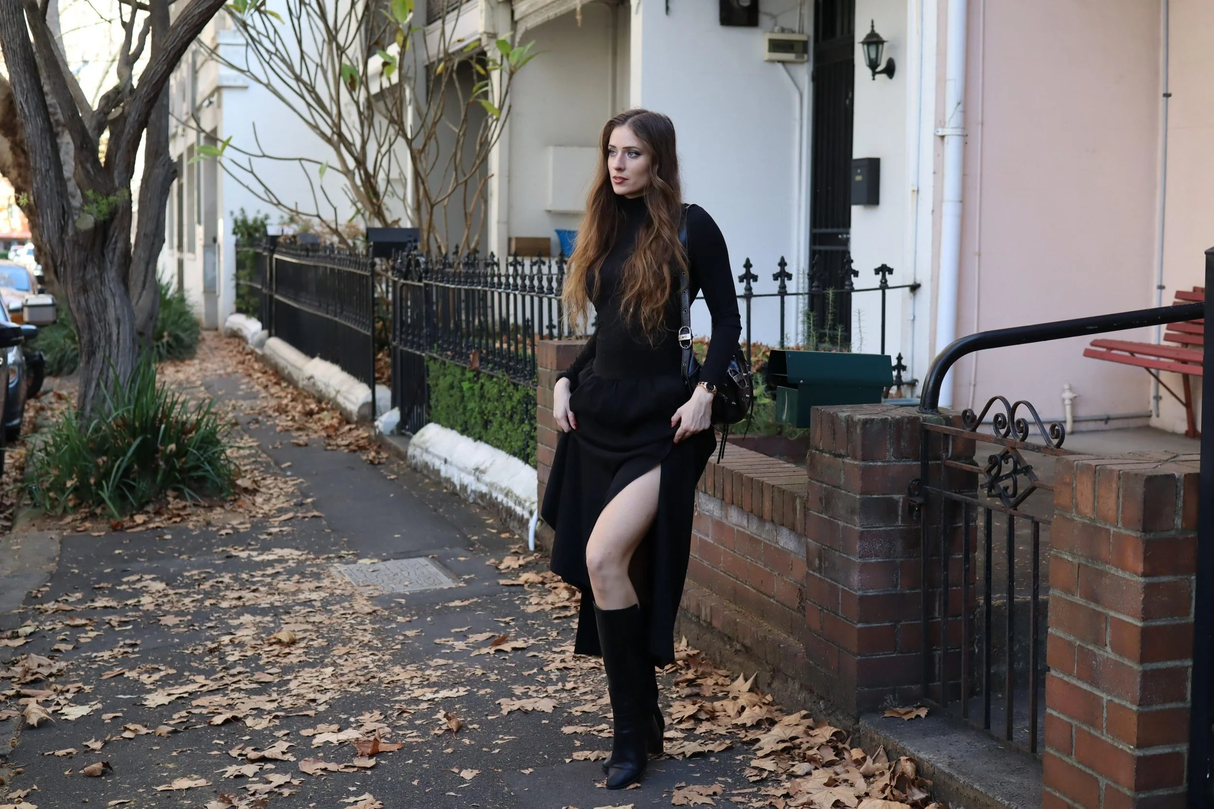A woman in a black dress with a high slit standing on a leaf-covered sidewalk next to a brick wall and iron gate, with houses and trees in the background.
