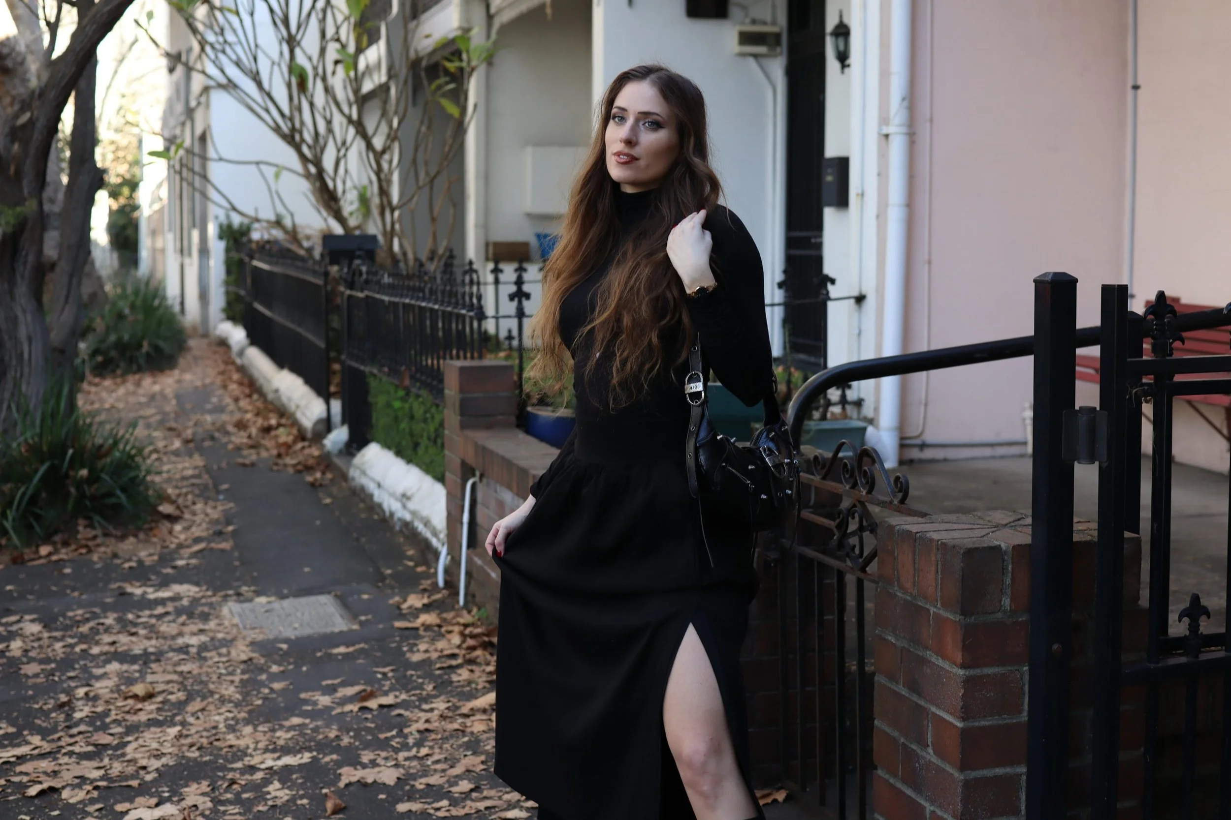A young woman with long brown hair in a black dress with a side slit, standing on a leaf-covered sidewalk, holding a black handbag, and posing in front of a residential fence.
