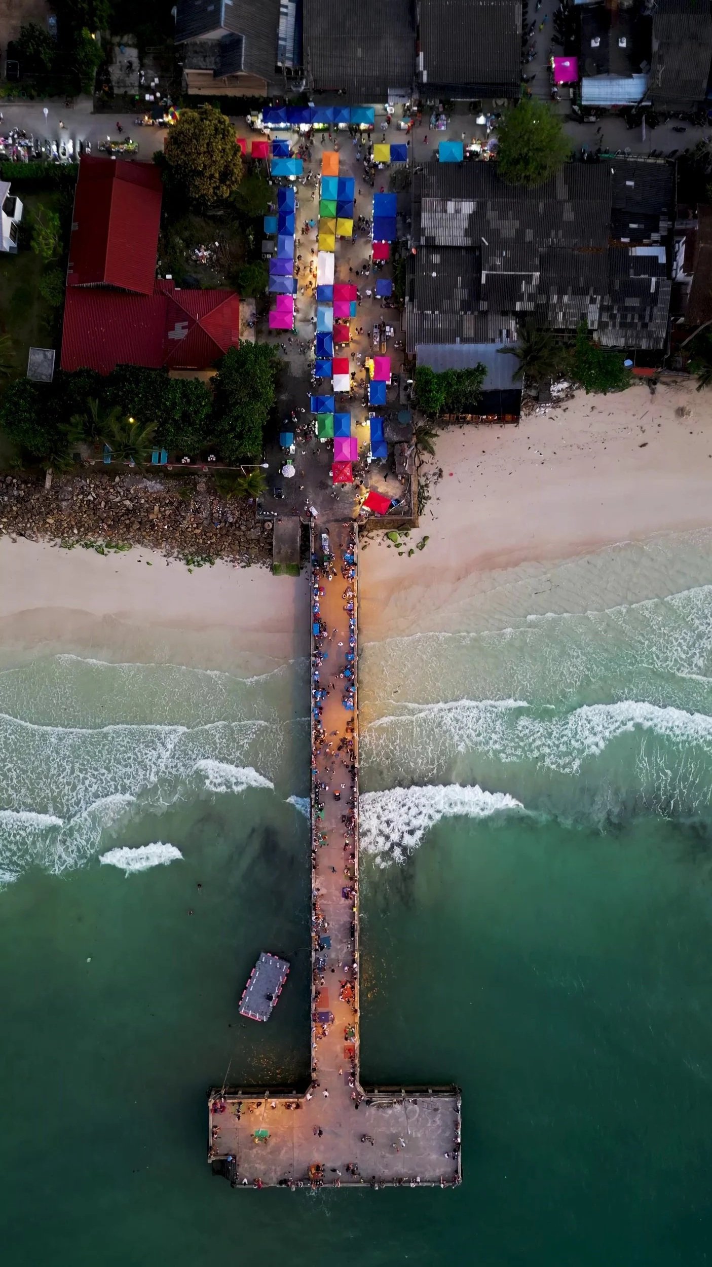 An aerial view of a beachfront pier extending into the ocean, with a sandy beach on one side and a lively market area with colorful tents on the other.
