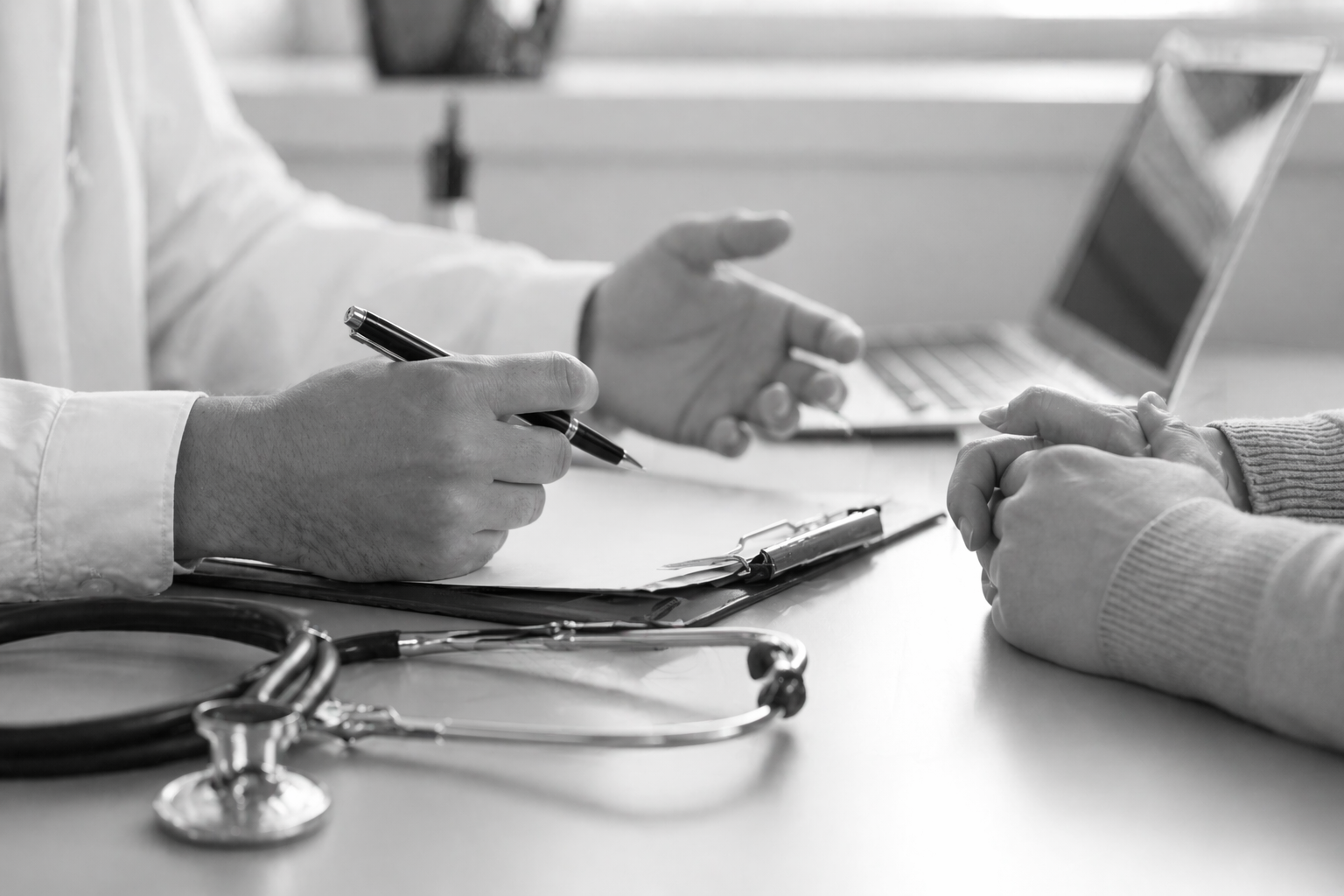 Black and white close-up photograph of a rheumatology consultation, showing a doctor’s hands with a pen and stethoscope on one side of the desk and the patient seated opposite, illustrating a second-opinion appointment.