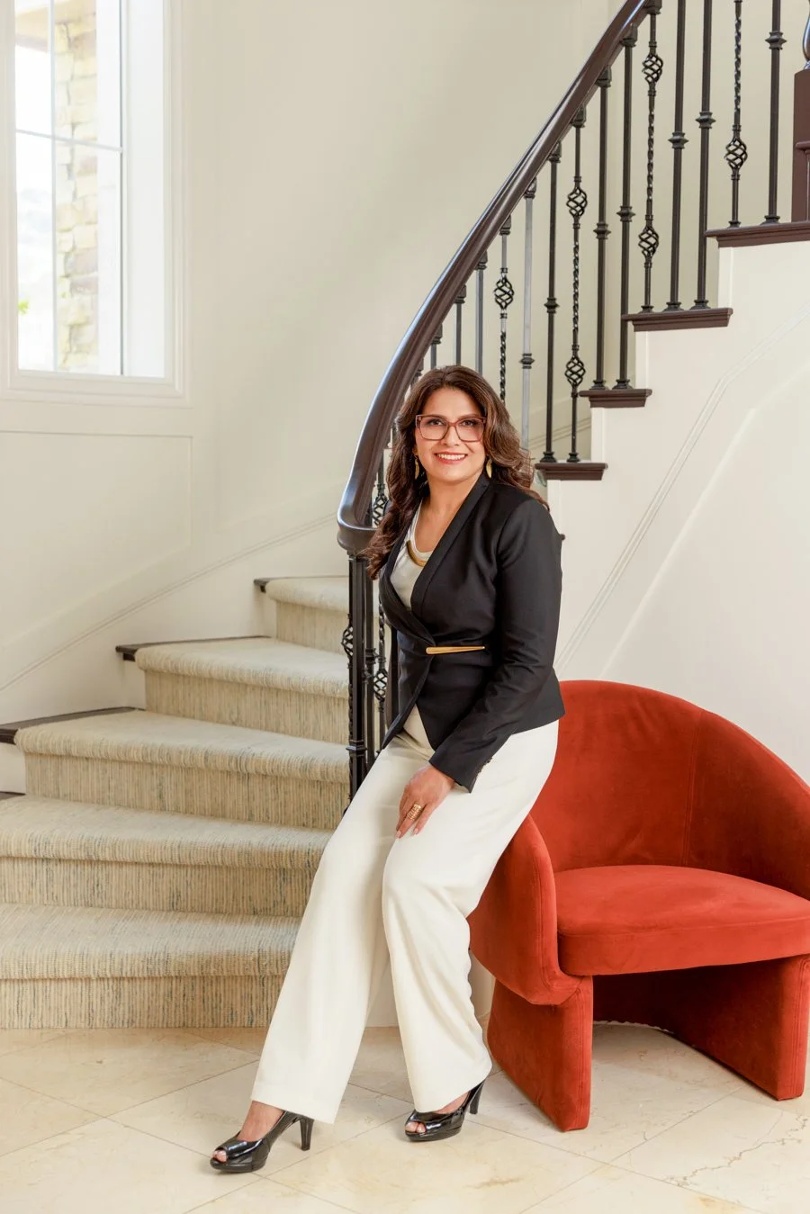 A woman in a black blazer, white pants, and high heels sitting on a red chair next to a staircase with black iron railing inside a house.