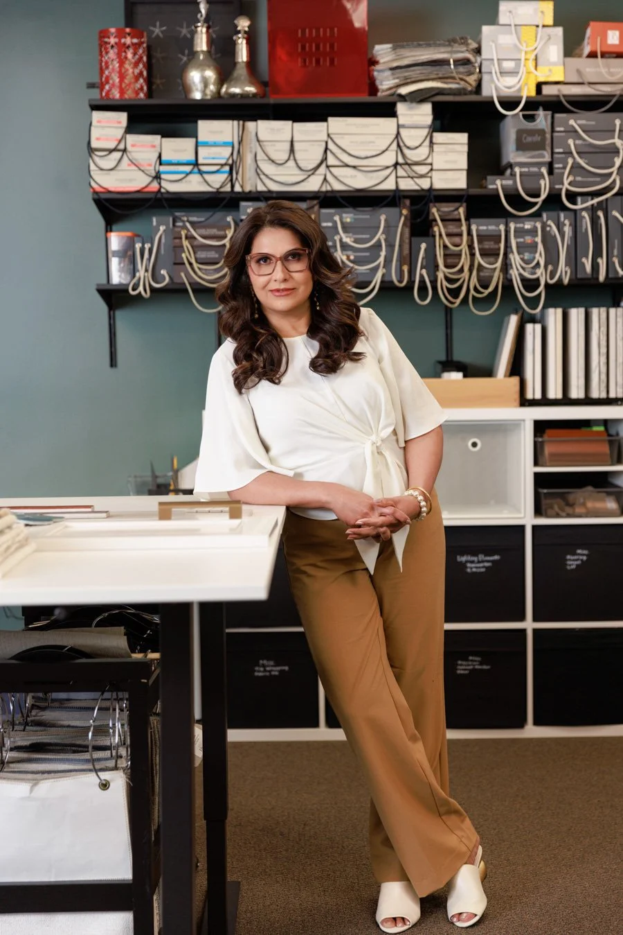 A woman with wavy brown hair and glasses standing in an office or design studio, leaning on a white table with a background of shelves containing boxes, books, and decorative items.
