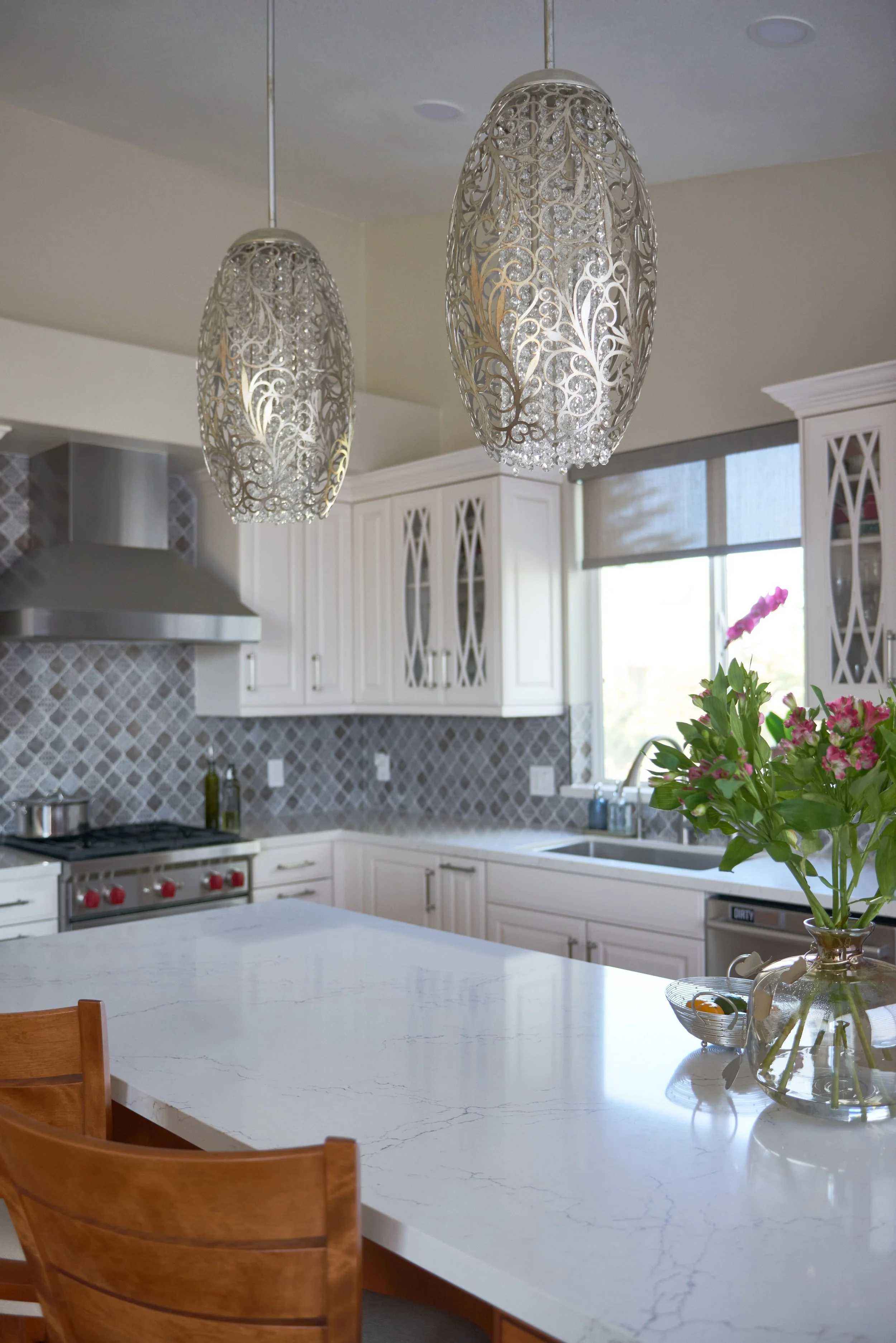 Modern kitchen with white cabinets, stainless steel appliances, decorative tile backsplash, and a white marble island. Two ornate pendant lights hang from the ceiling, and a vase with pink flowers is on the counter.