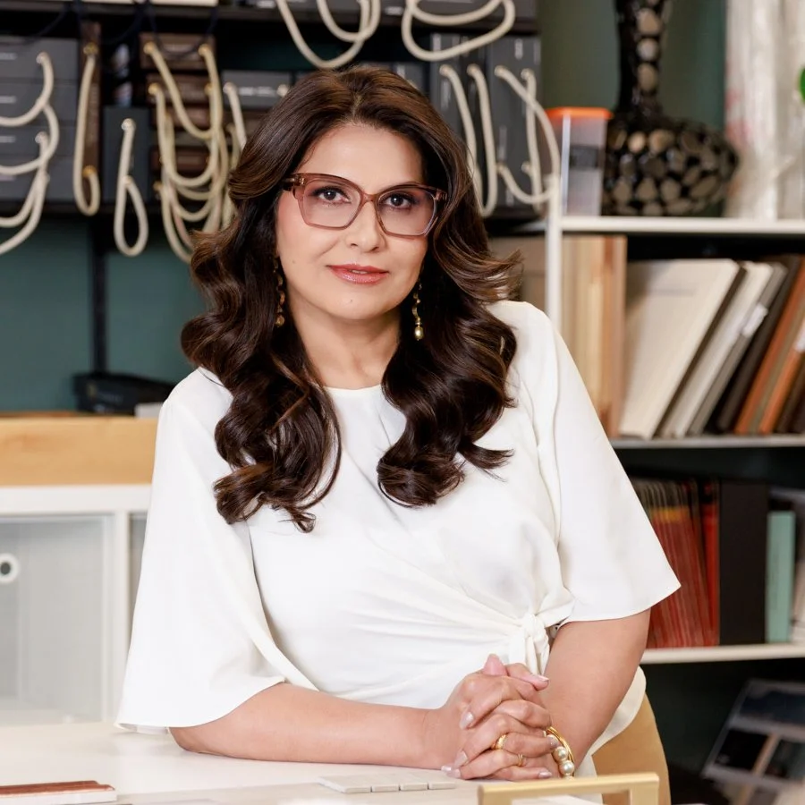 A woman with long, wavy brown hair, wearing glasses and earrings, sitting at a desk in an office or library with bookshelves and decorative wires in the background.